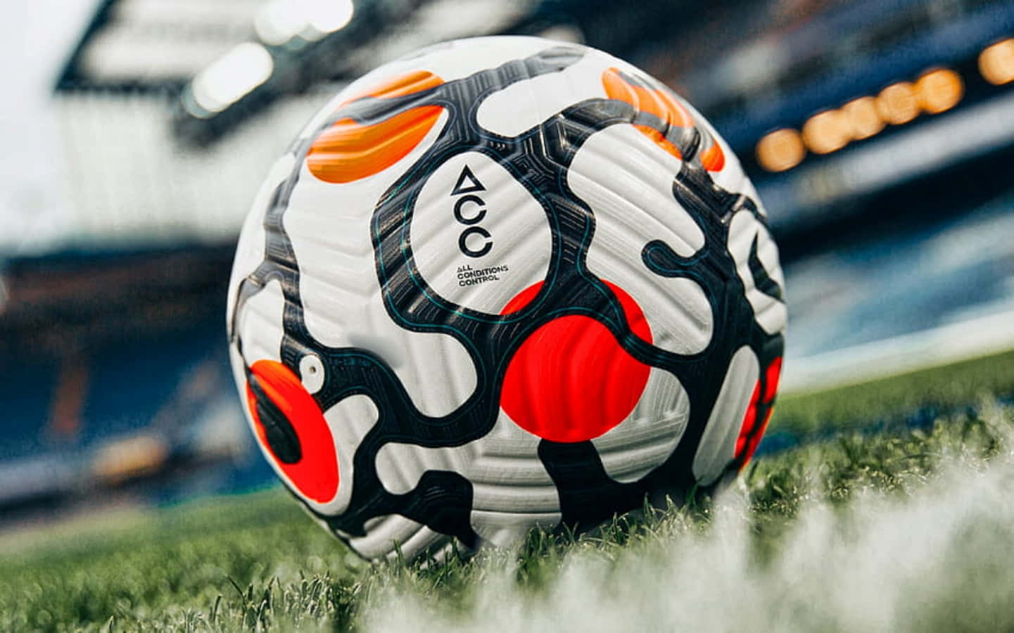 Soccer Ball 桌布 壁紙 — 一個白色、黑色和橙色的足球特寫，放在充滿活力的綠色草地上，背景有球場燈光閃爍。