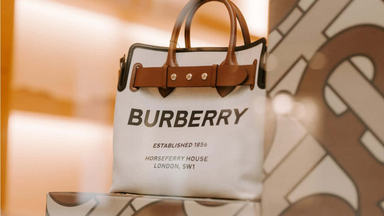 Burberry 桌布 壁紙 — 一個白色Burberry手提包搭配棕色皮革肩帶，優雅地置於溫暖光線下的模糊圖案背景前。