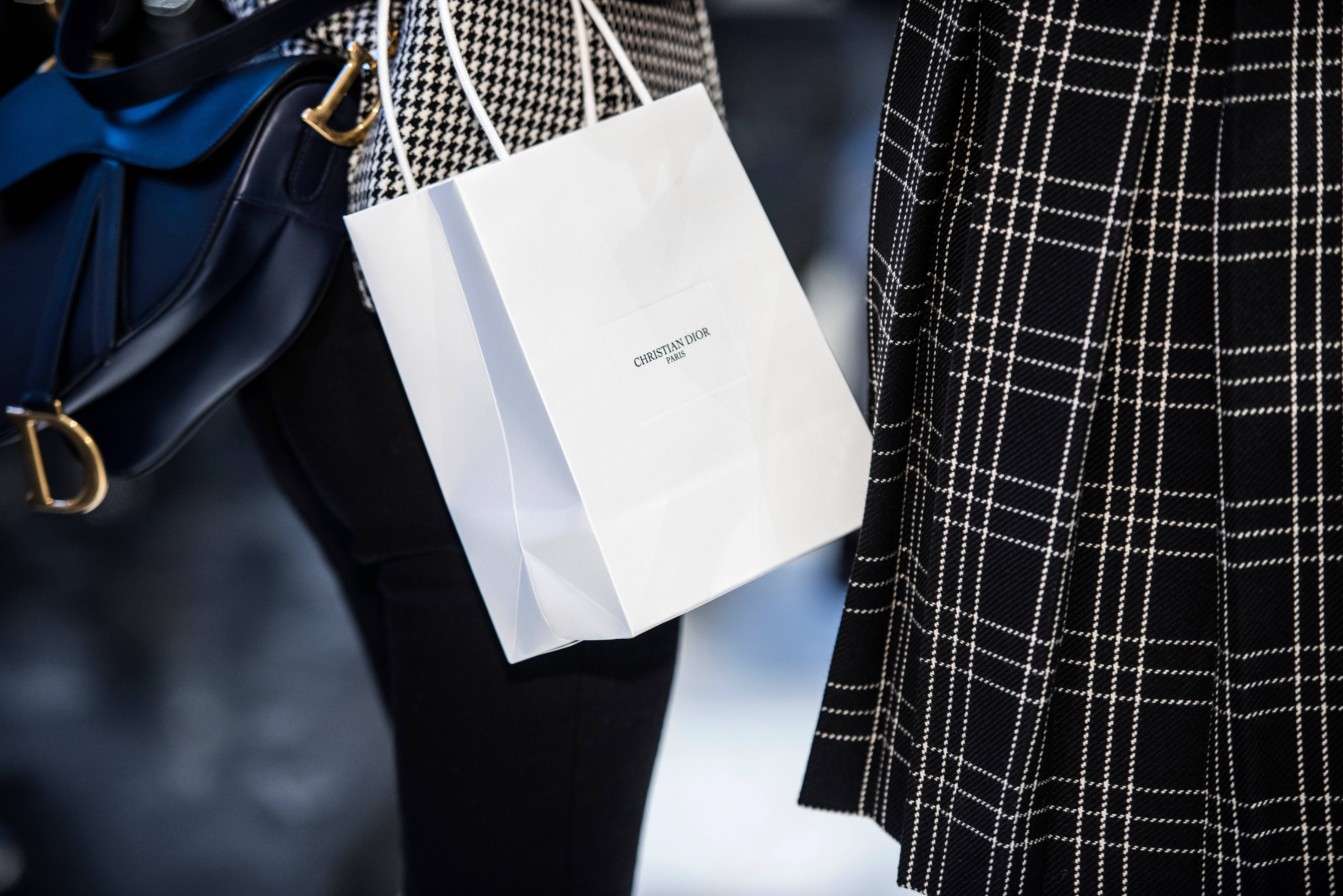 Christian Dior 桌布 壁紙 — 一個白色Christian Dior購物袋的特寫，由一個穿著黑白格紋裝束並拿著藍色皮手袋的人拿著。