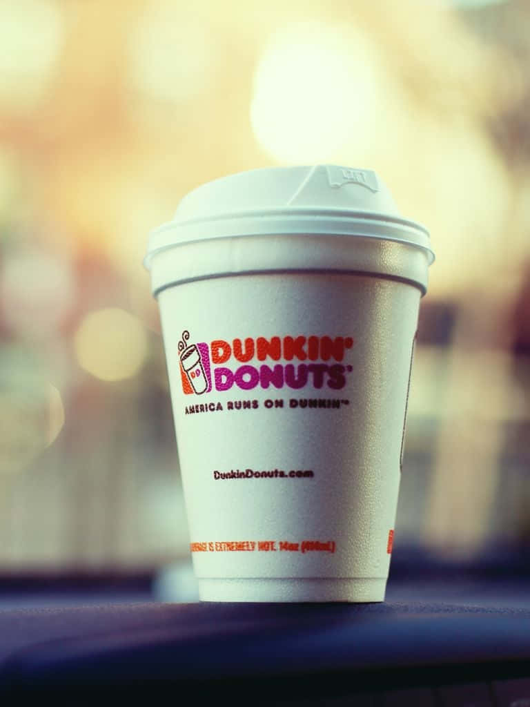 Dunkin Donuts 桌布 壁紙 — 一個白色Dunkin' Donuts咖啡杯連蓋，放在深色表面上，背景溫暖模糊。
