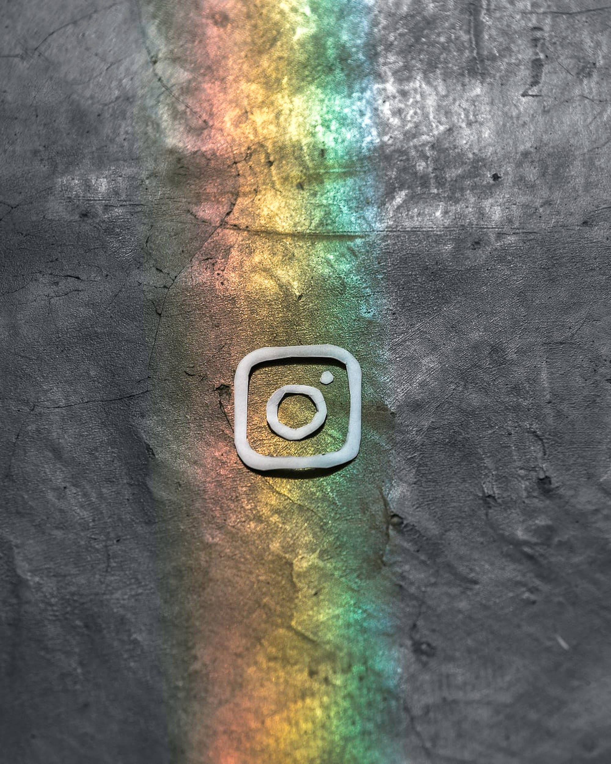 Instagram 桌布 壁紙 — 一個白色Instagram標誌放在一個破裂的灰色表面上，被一條充滿活力的彩虹光條照亮。