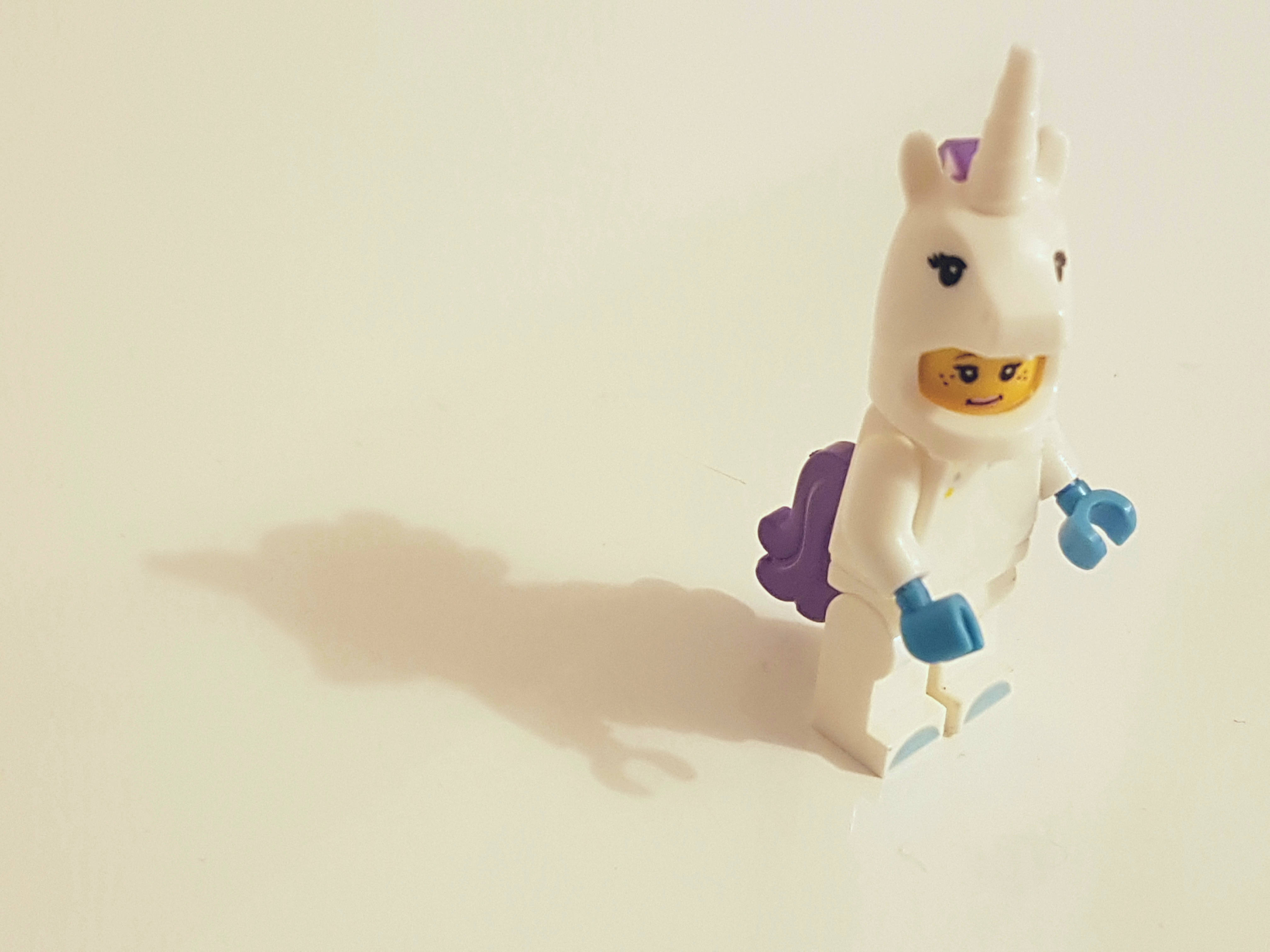 獨角獸 桌布 壁紙 — 一個白色LEGO迷你人偶打扮成獨角獸，有紫色鬃毛和藍色手，站在純白表面，投下柔和陰影。
