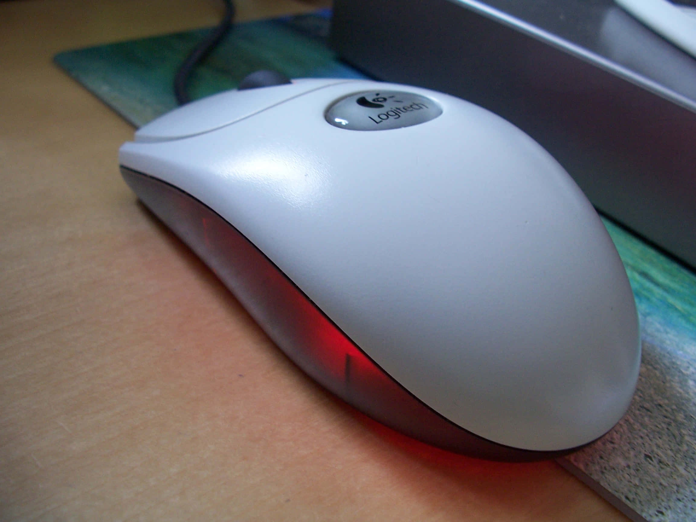 Device 桌布 壁紙 — 一個白色Logitech電腦滑鼠，底部發出紅色光芒，放在木桌上，旁邊有一個彩色滑鼠墊。