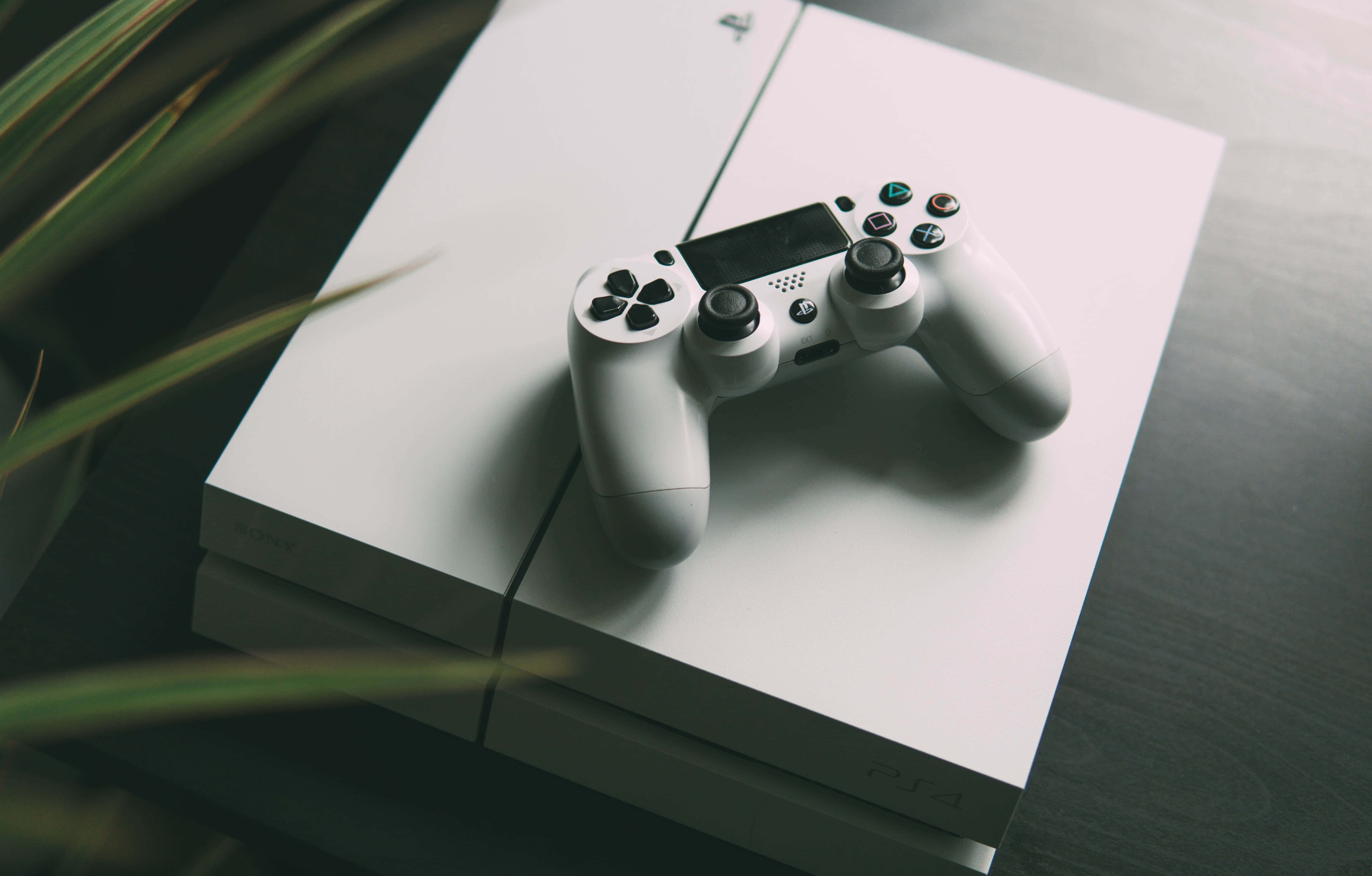Gaming Consoles 桌布 壁紙 — 一個白色PlayStation 4手把放在主機上，柔和自然光線，前景有模糊的綠色植物葉子。