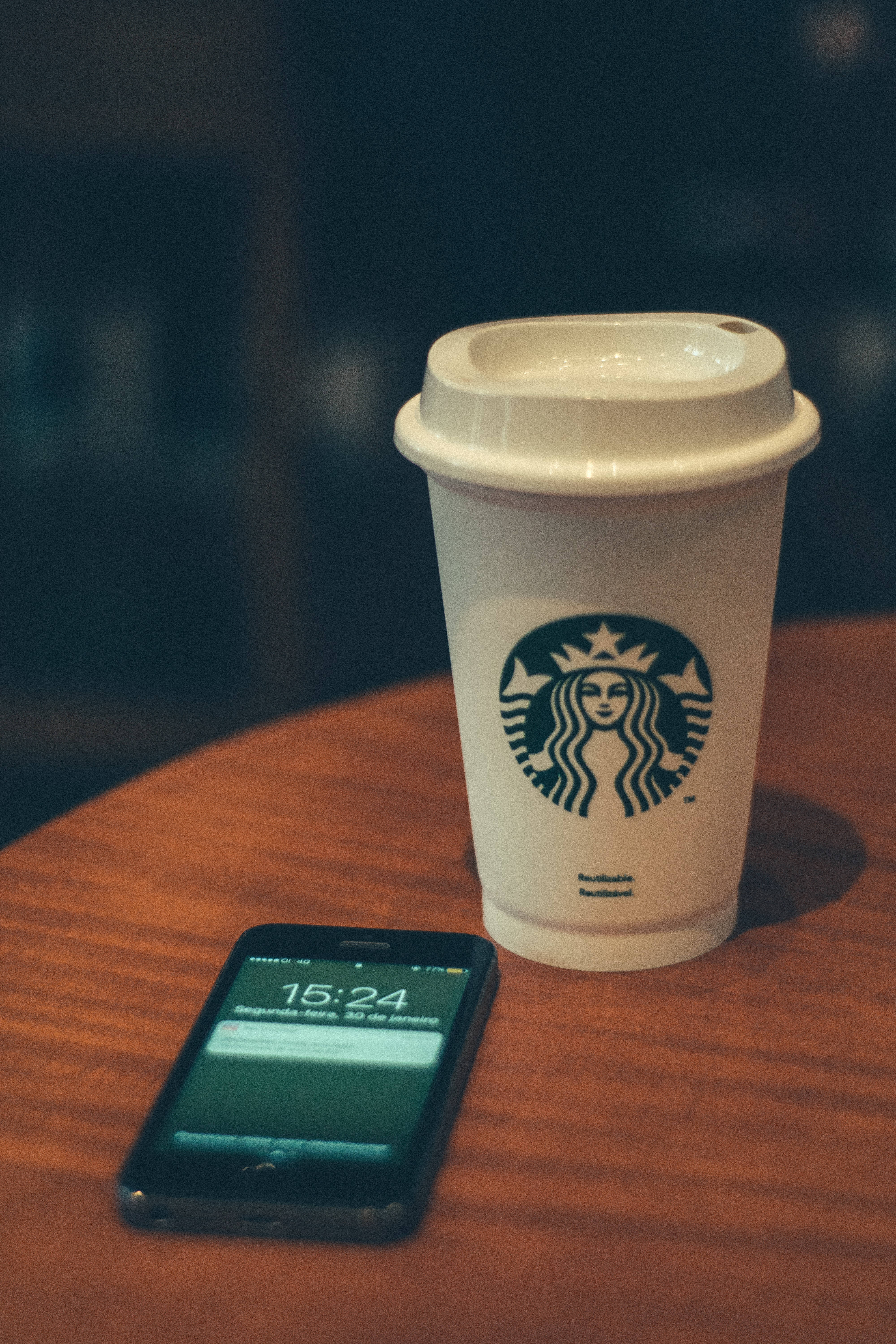 蘋果 桌布 壁紙 — 一個白色Starbucks杯和黑色智慧型手機放在溫暖的木桌上，背景是柔和模糊的咖啡館環境。