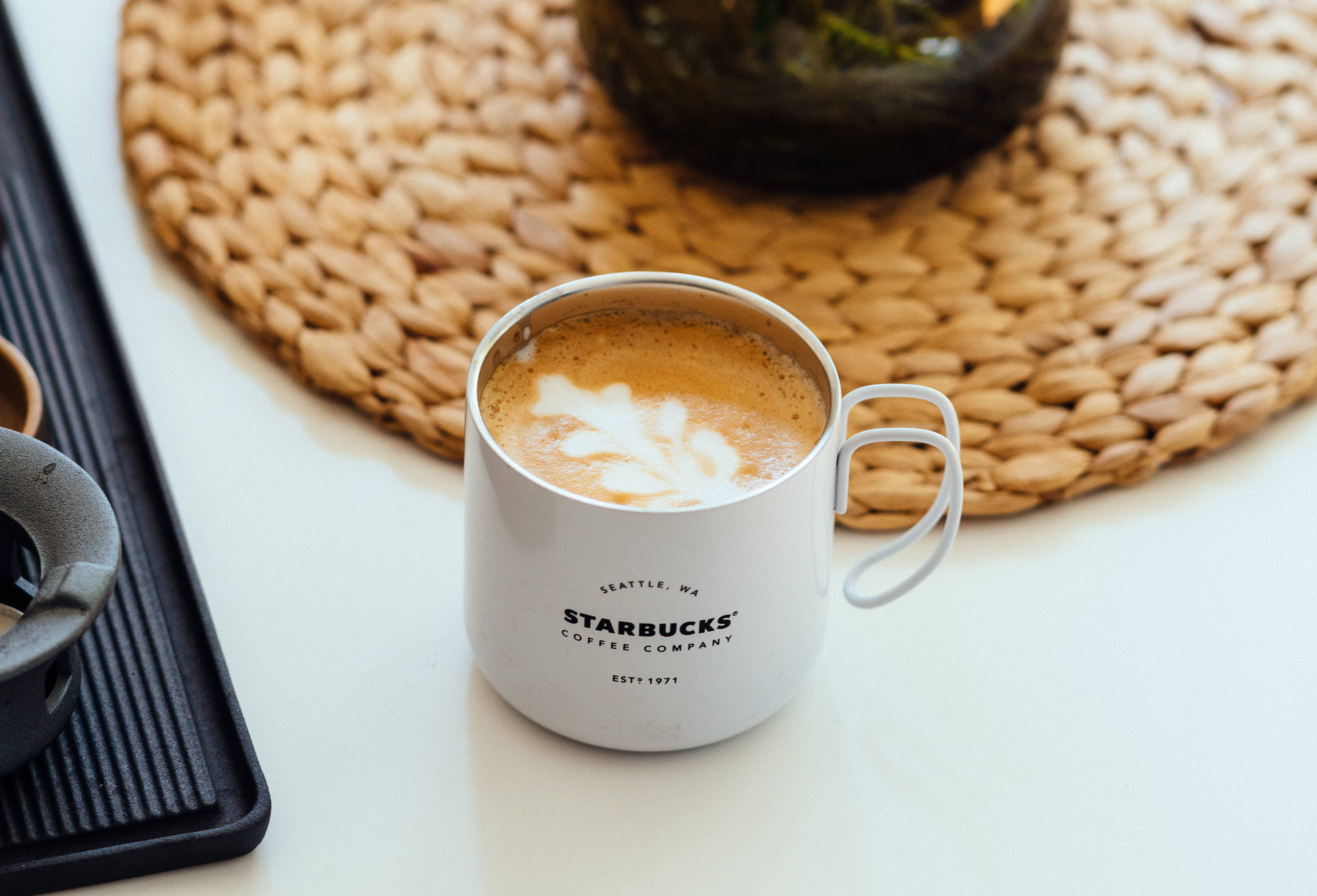 星巴克 桌布 壁紙 — 一個白色Starbucks馬克杯，上面有葉子拉花藝術，放在編織墊上，旁邊有一個黑色托盤和玻璃罐。