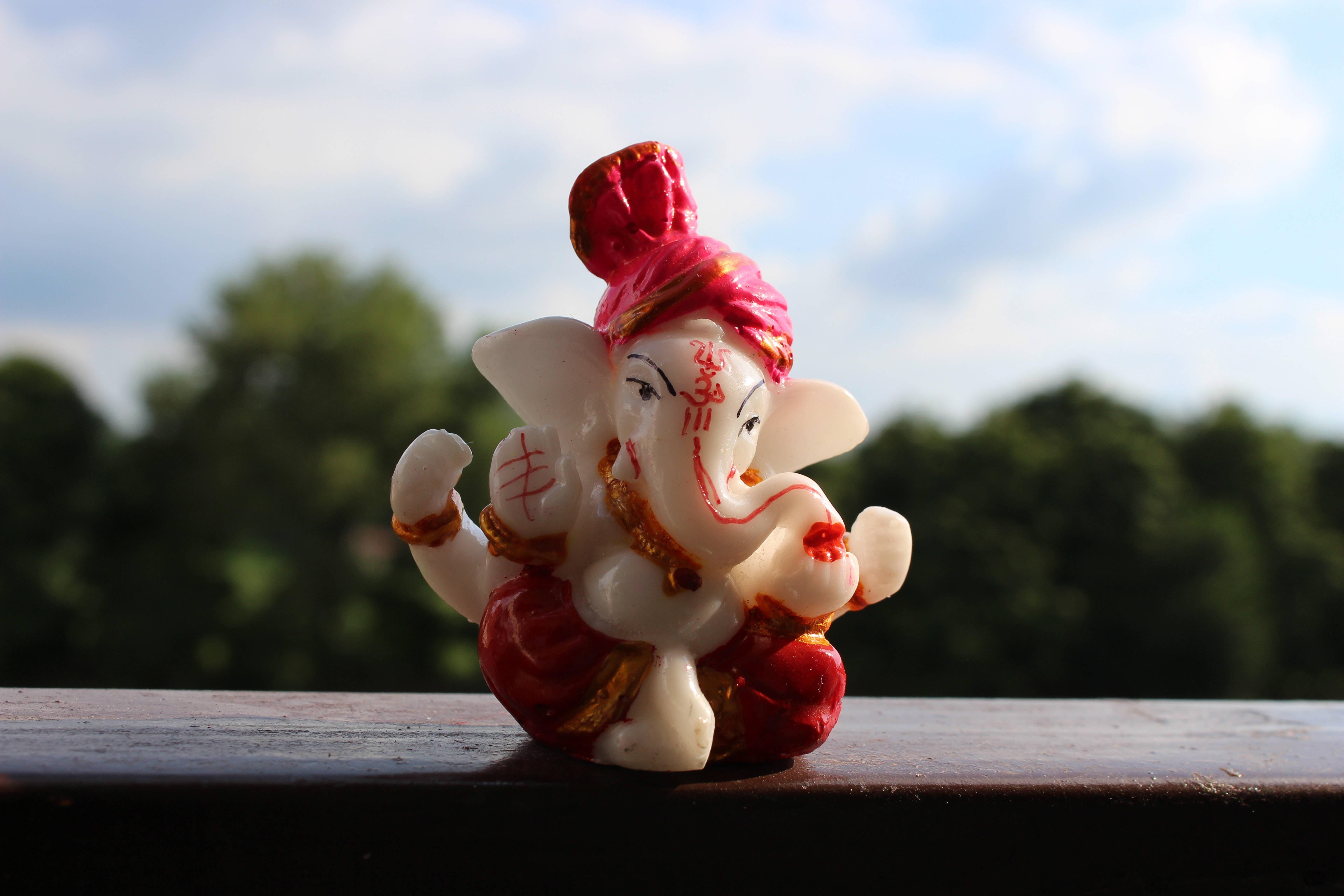 ganesh 4K 桌布 壁紙 — 一個白色和紅色的象神雕像坐在木欄杆上，背景是模糊的綠色樹木和藍天。
