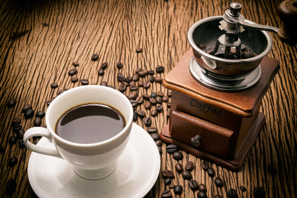 Brown Coffee 桌布 壁紙 — 一個白色咖啡杯和一個復古木製研磨機放在一個散落著烘焙咖啡豆的質樸木桌上。