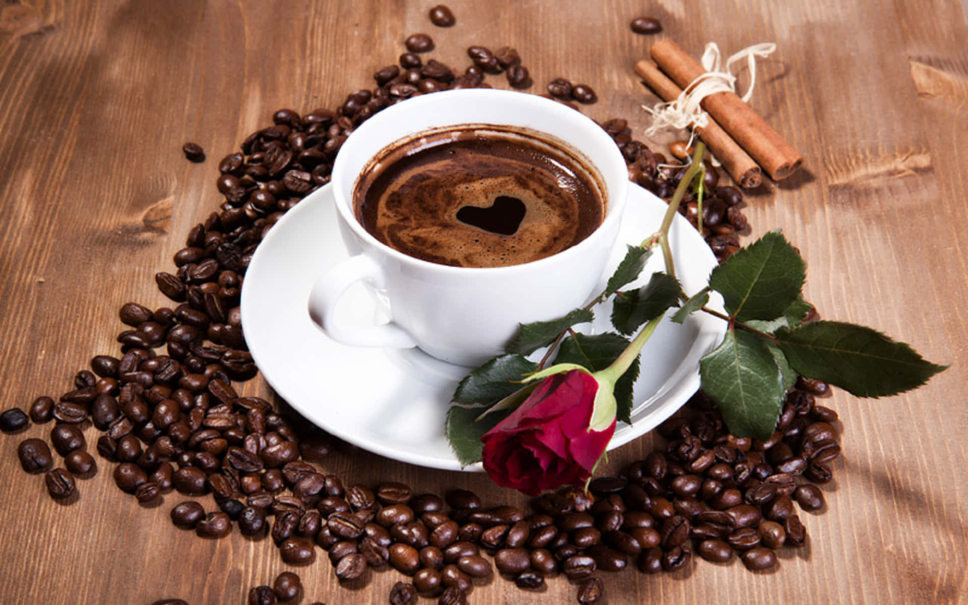 Morning Coffee 桌布 壁紙 — 一個白色咖啡杯帶有心形奶泡，放在木桌上，周圍有咖啡豆、紅玫瑰和肉桂棒。