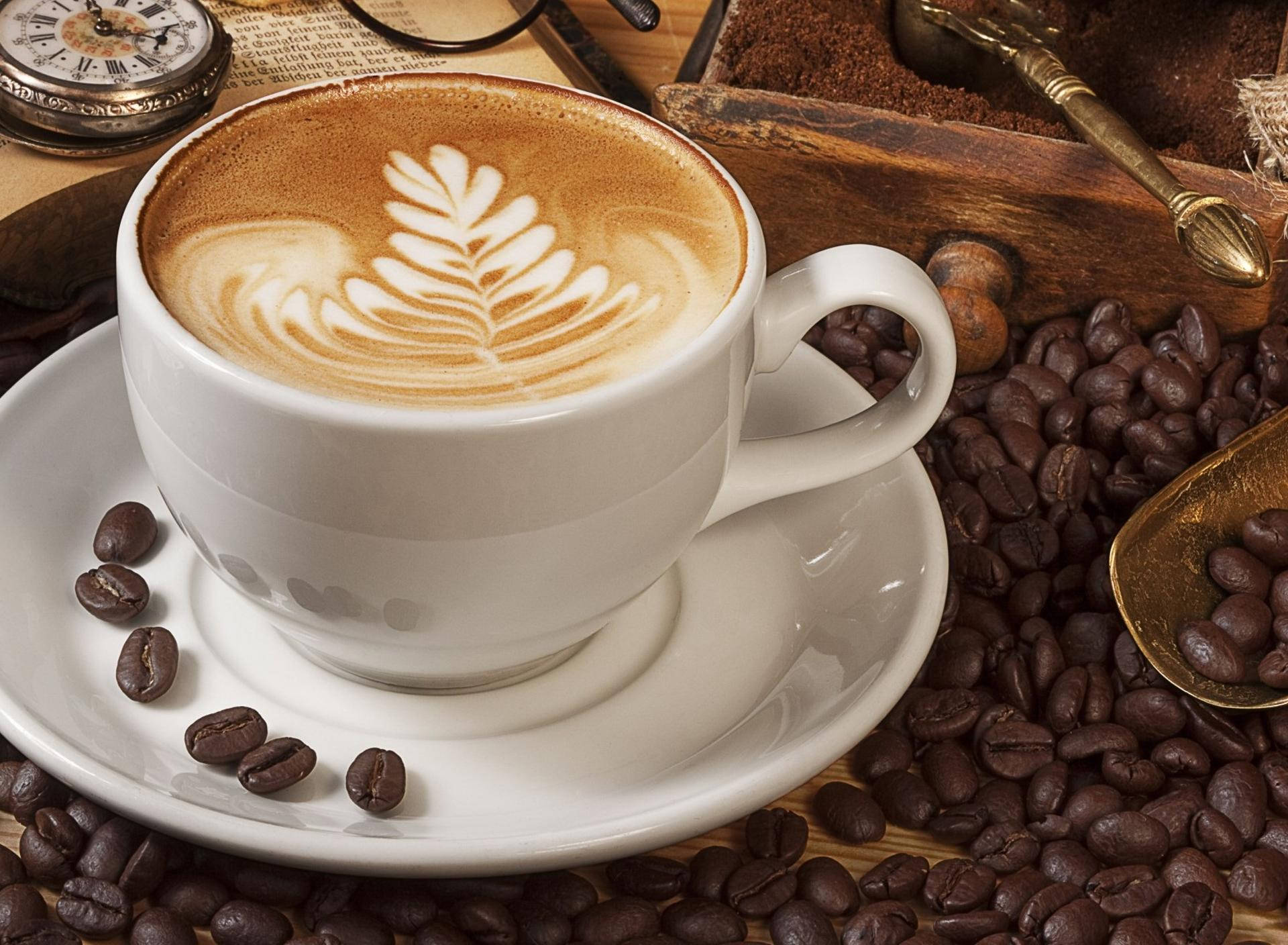 Coffee Aesthetic 桌布 壁紙 — 一個白色咖啡杯帶有葉子拉花藝術，放在碟子上，周圍環繞著烘焙咖啡豆和復古道具。