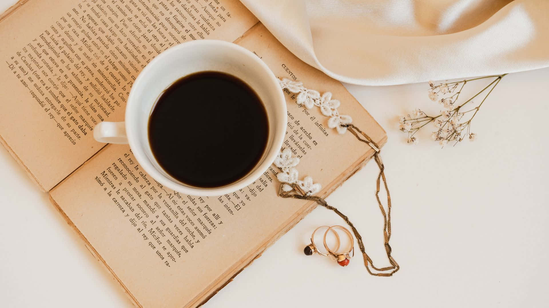 Brown Pastel Aesthetic 桌布 壁紙 — 一個白色咖啡杯放在一本打開的書上，旁邊有乾燥花和戒指在柔軟的奶油色布料上。