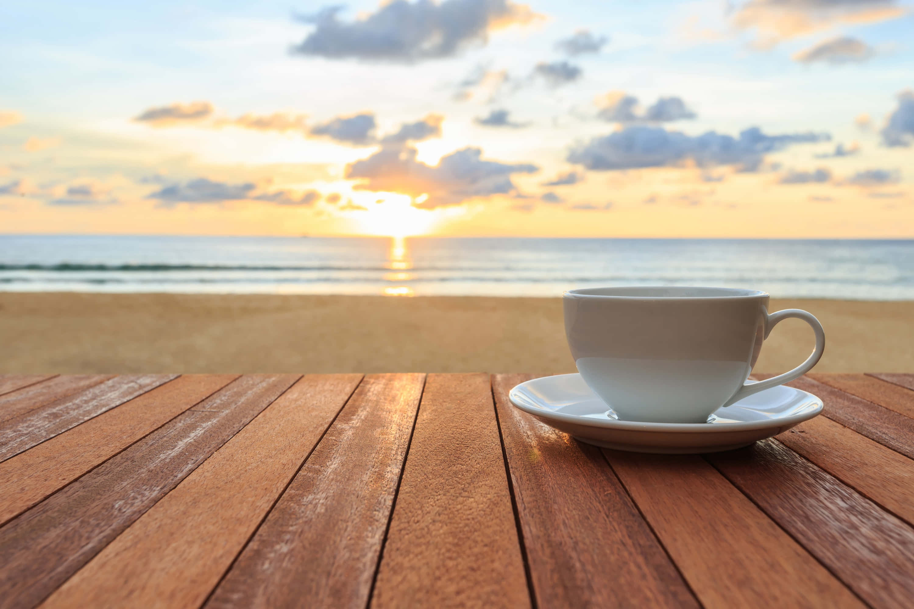 Morning Coffee 桌布 壁紙 — 一個白色咖啡杯放在木桌上，俯瞰日落時分寧靜的海灘，金色雲彩和平靜的海浪。