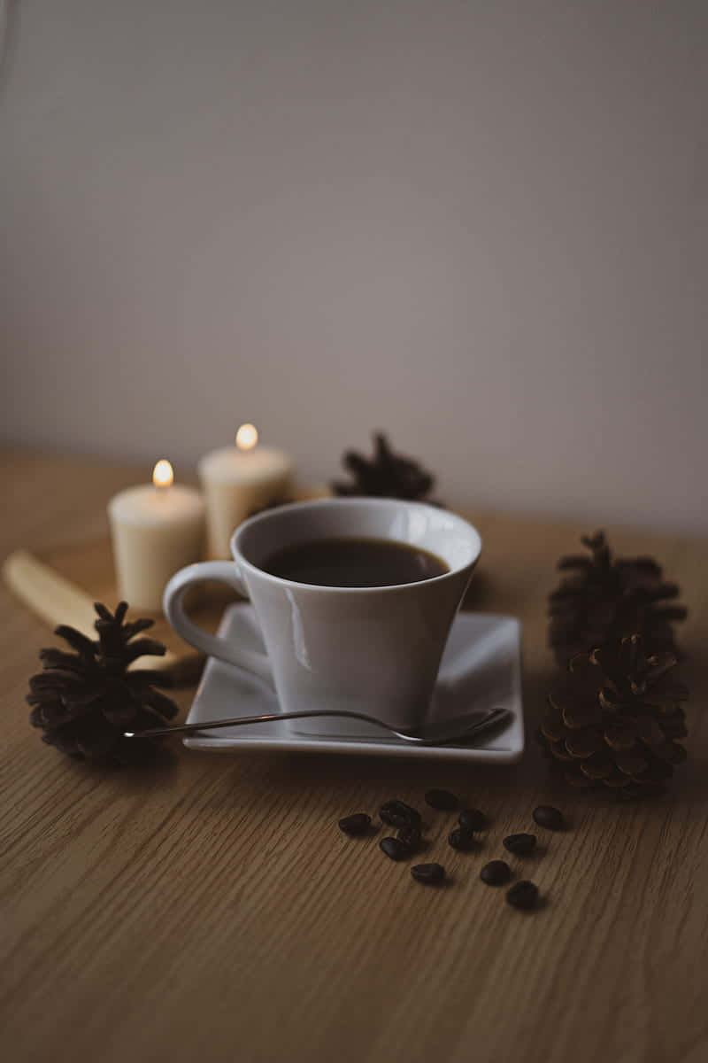 Coffee Aesthetic 桌布 壁紙 — 一個白色咖啡杯放在木桌上，旁邊有松果和點燃的蠟燭，營造出舒適的冬季氛圍。