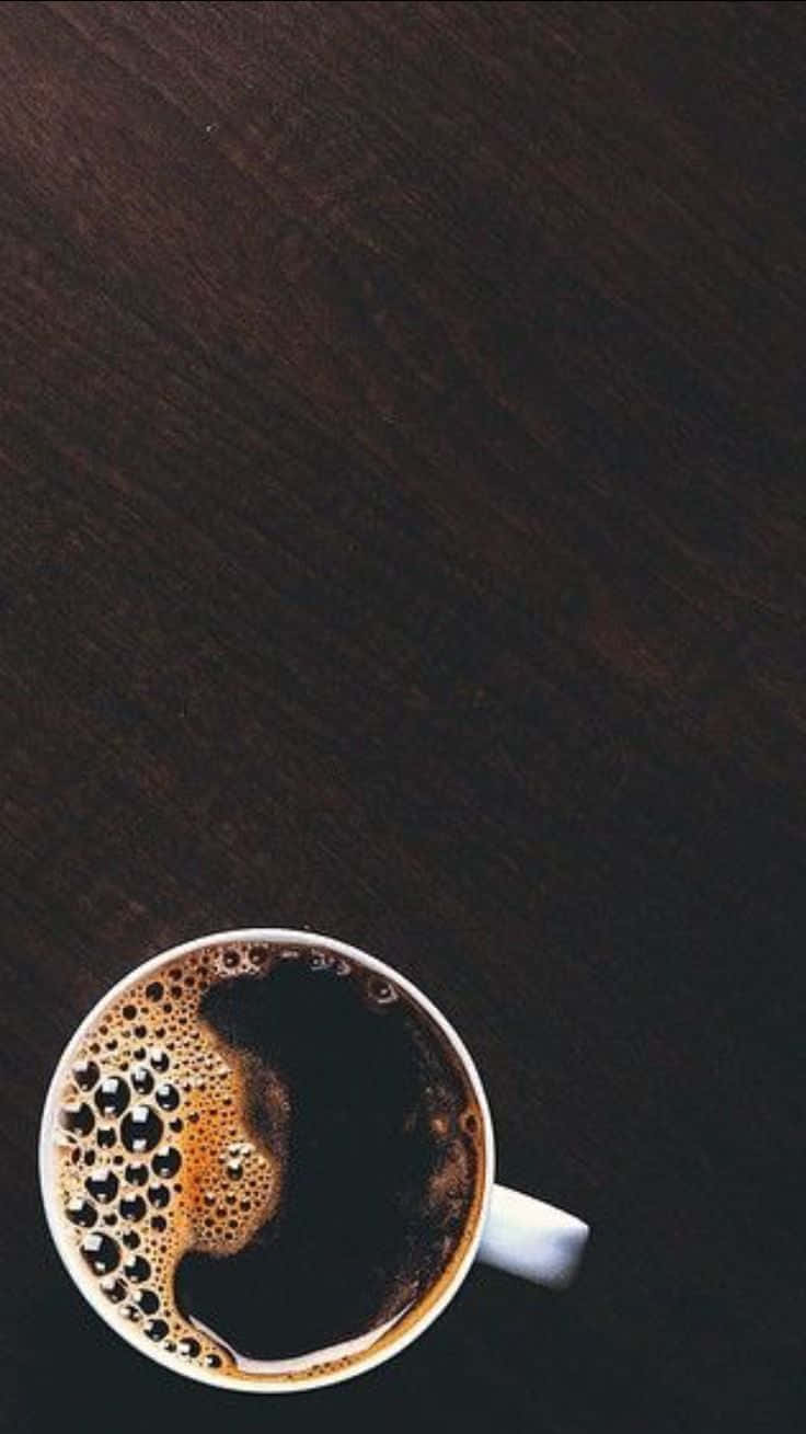 Black Coffee 桌布 壁紙 — 一個白色咖啡杯裝滿深色咖啡和泡沫，放在從上方俯瞰的深色木桌上。