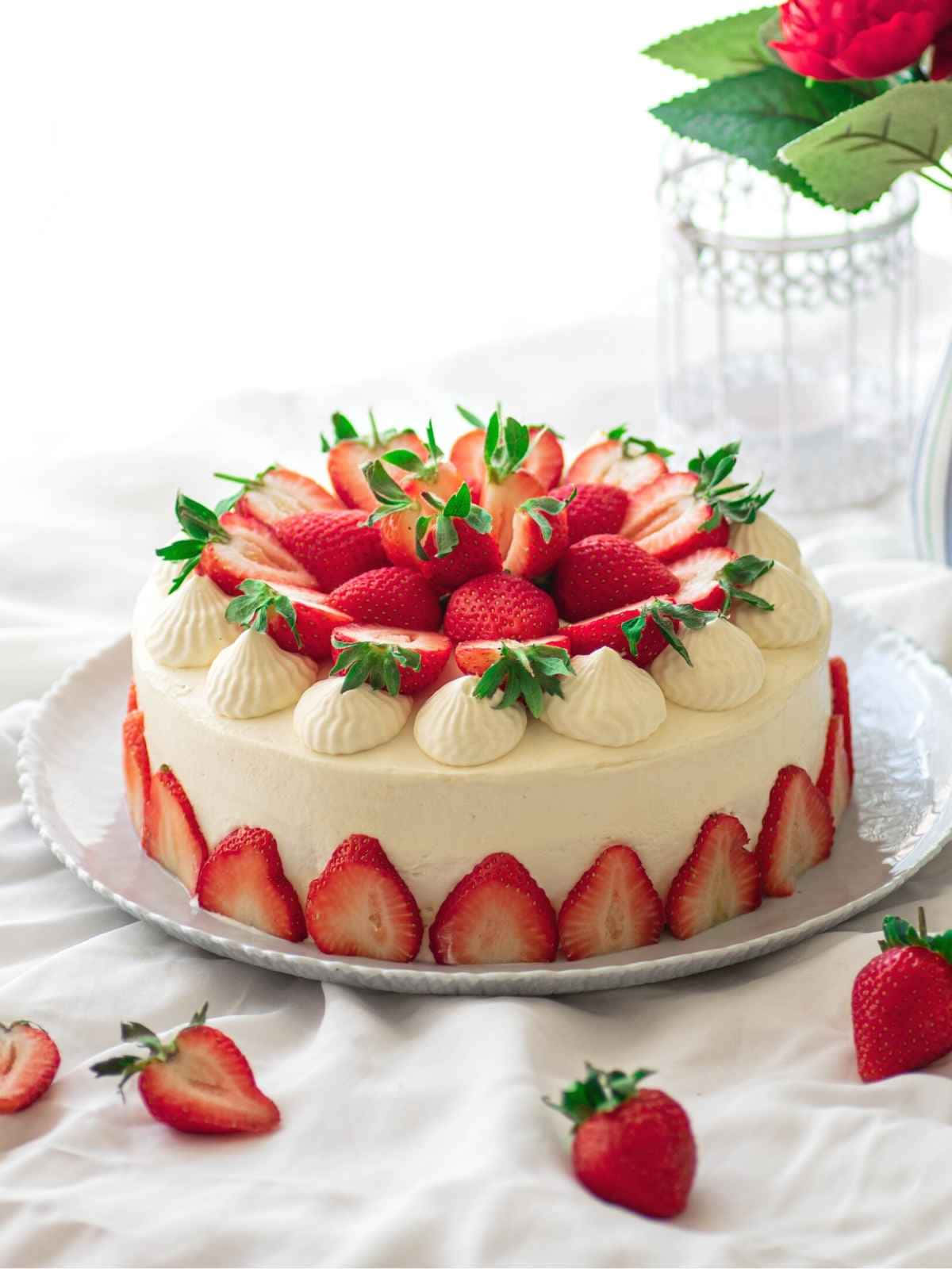Strawberries 桌布 壁紙 — 一個白色圓形蛋糕，飾以新鮮草莓和奶油玫瑰花，放在白色盤子上，背景有散落的草莓和一朵紅玫瑰。