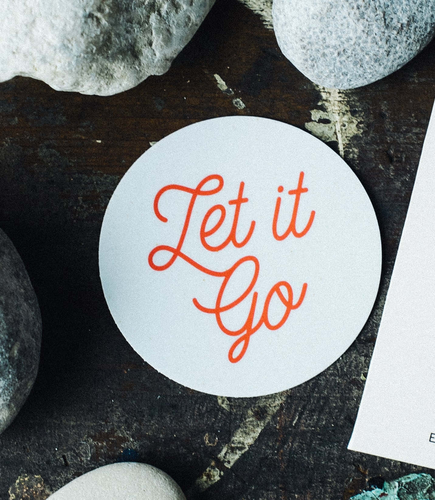 語錄 桌布 壁紙 — 一個白色圓形貼紙，上面有橙色草書文字寫著「Let it Go」，放在深色紋理表面的灰色河石之間。