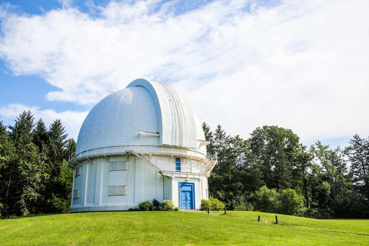 Observatory 桌布 壁紙 — 一個白色天文台圓頂，帶有藍色門，位於長滿青草的山丘上，周圍是茂密的綠樹，天空部分多雲。