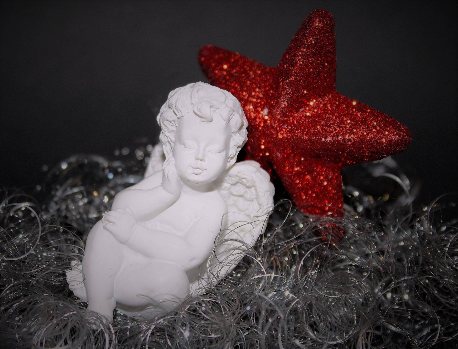 angels 桌布 壁紙 — 一個白色小天使雕像在黑暗背景下，安詳地躺在閃爍的紅星旁邊，周圍是銀色閃粉。