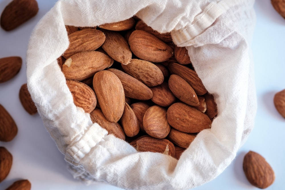 Almond 桌布 壁紙 — 一個白色布袋裝滿棕色杏仁，溢出到明亮的白色表面上。