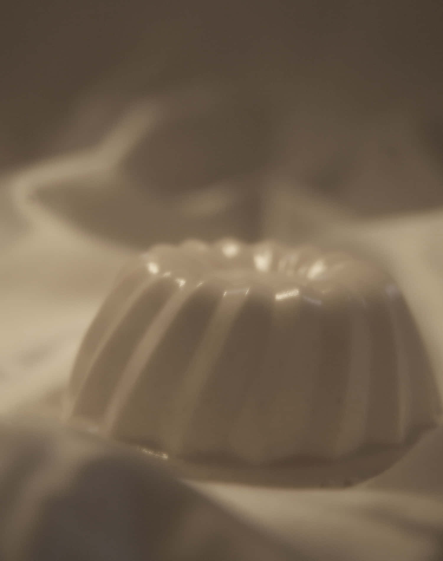 Blancmange 桌布 壁紙 — 一個白色帶凹槽的甜點模具特寫，放置在柔軟布料上，周圍有溫暖的環境光線。