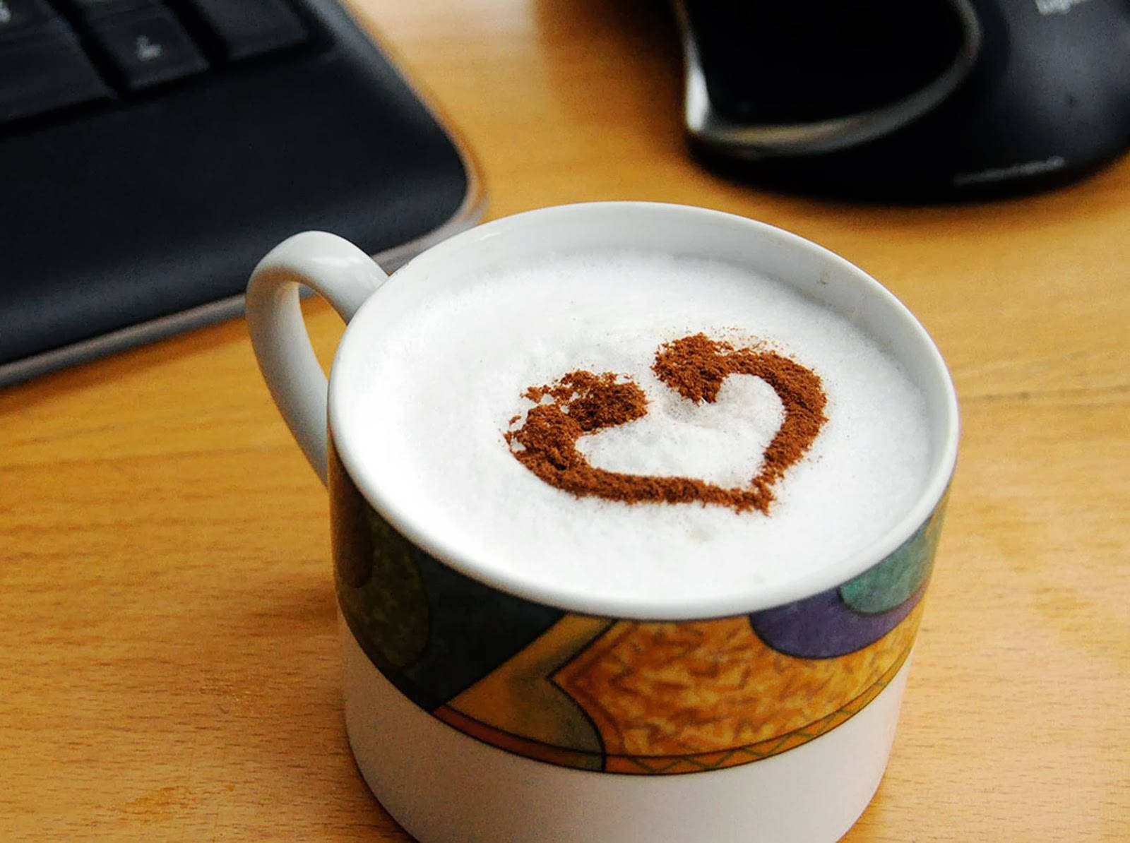 Coffee Aesthetic 桌布 壁紙 — 一個白色帶彩色圖案的馬克杯，裝著泡沫咖啡，上面放著肉桂心形裝飾，放在木桌上，旁邊有鍵盤和滑鼠。