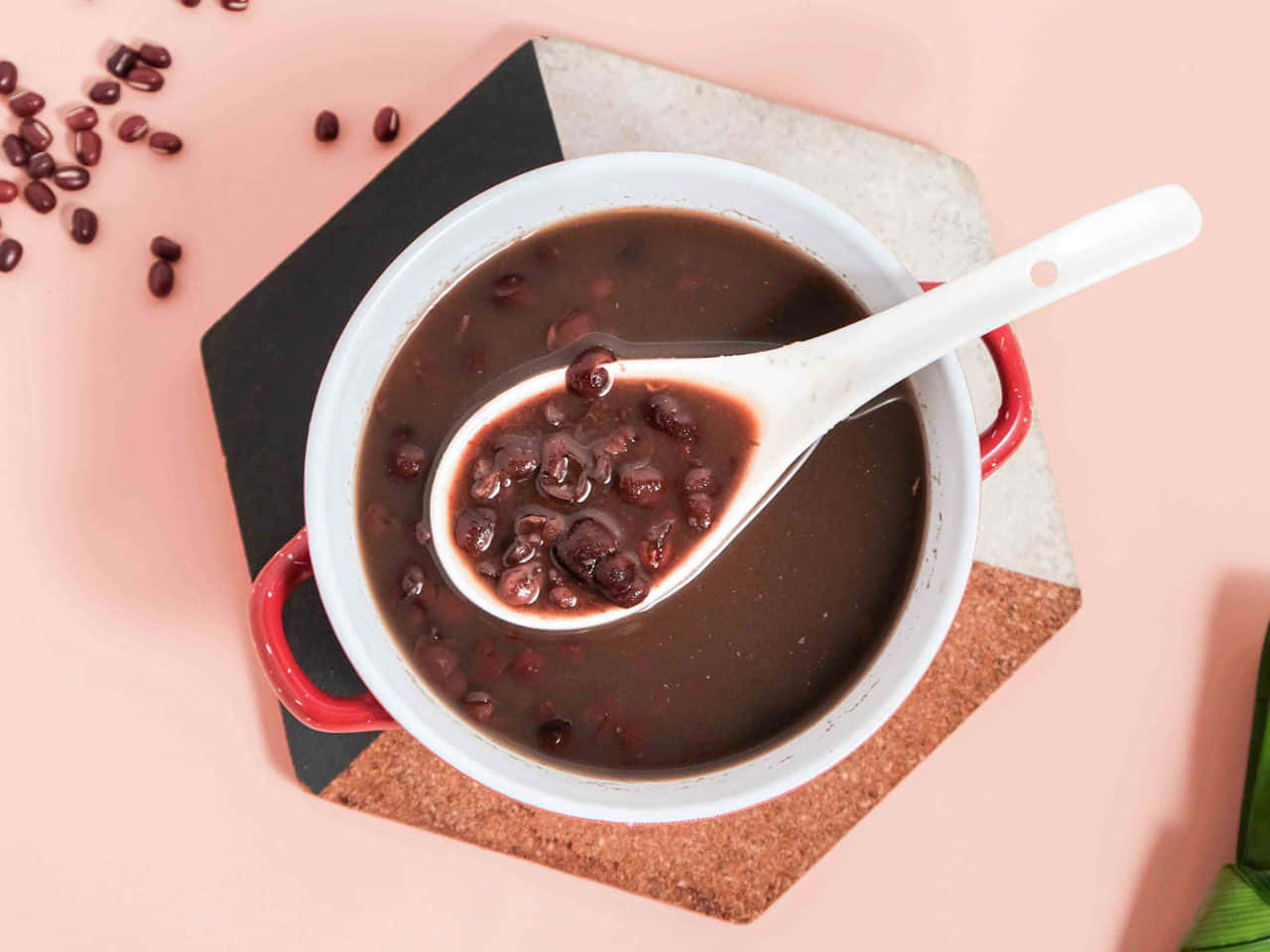 Red Beans 桌布 壁紙 — 一個白色帶紅色手柄的鍋盛載著濃郁的深紅豆湯，湯匙舀起光澤的豆子，襯托著柔和的粉紅色背景。