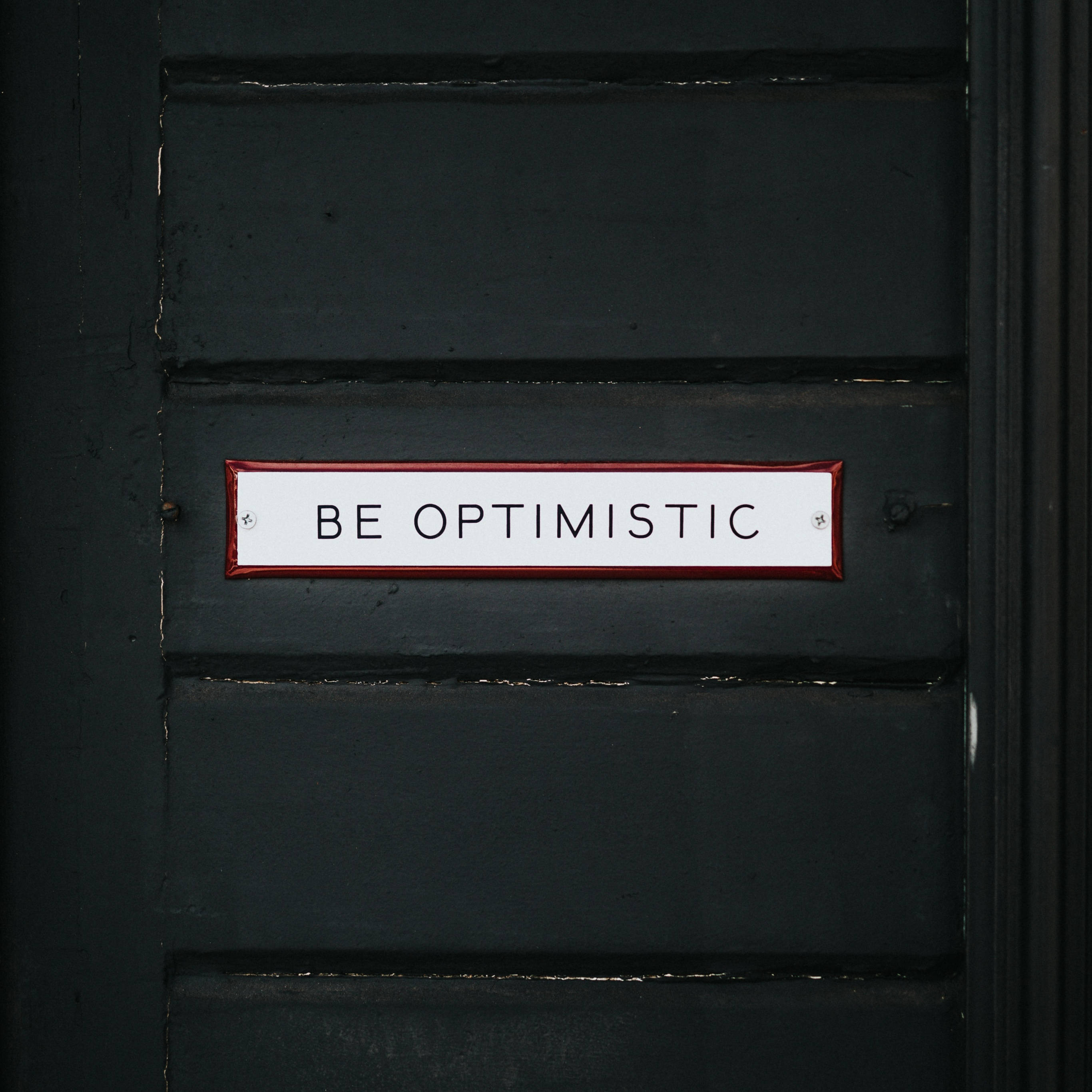 黑色 桌布 壁紙 — 一個白色帶紅色邊框的標誌，上面寫著 BE OPTIMISTIC，安裝在帶有可見木紋和螺絲的深綠色木門上。