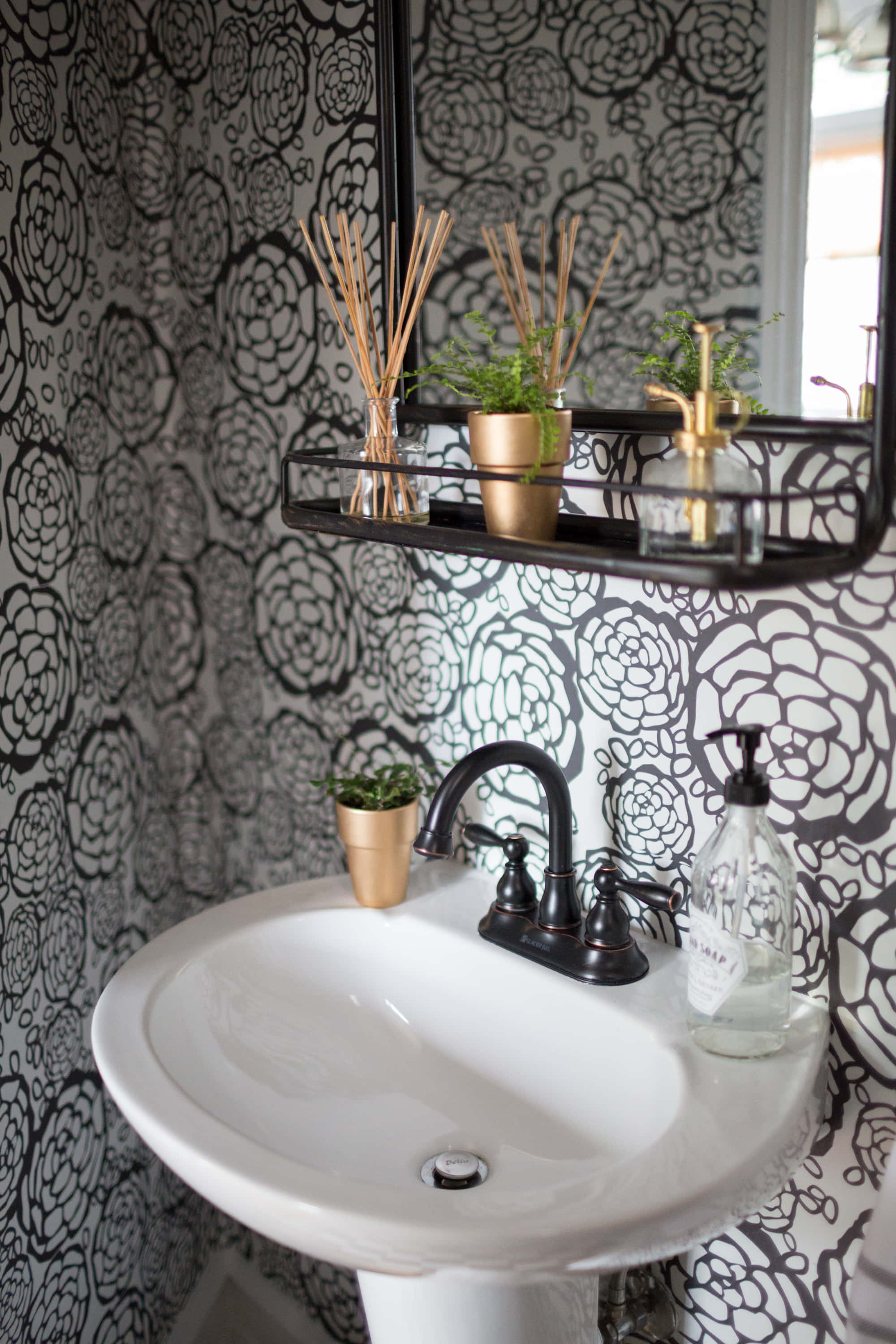 Bathroom Decor 桌布 壁紙 — 一個白色座地洗手盆搭配黑色水龍頭，放在大膽的黑白花卉壁紙前，黑色層架上的金色花盆和蘆葦作點綴。