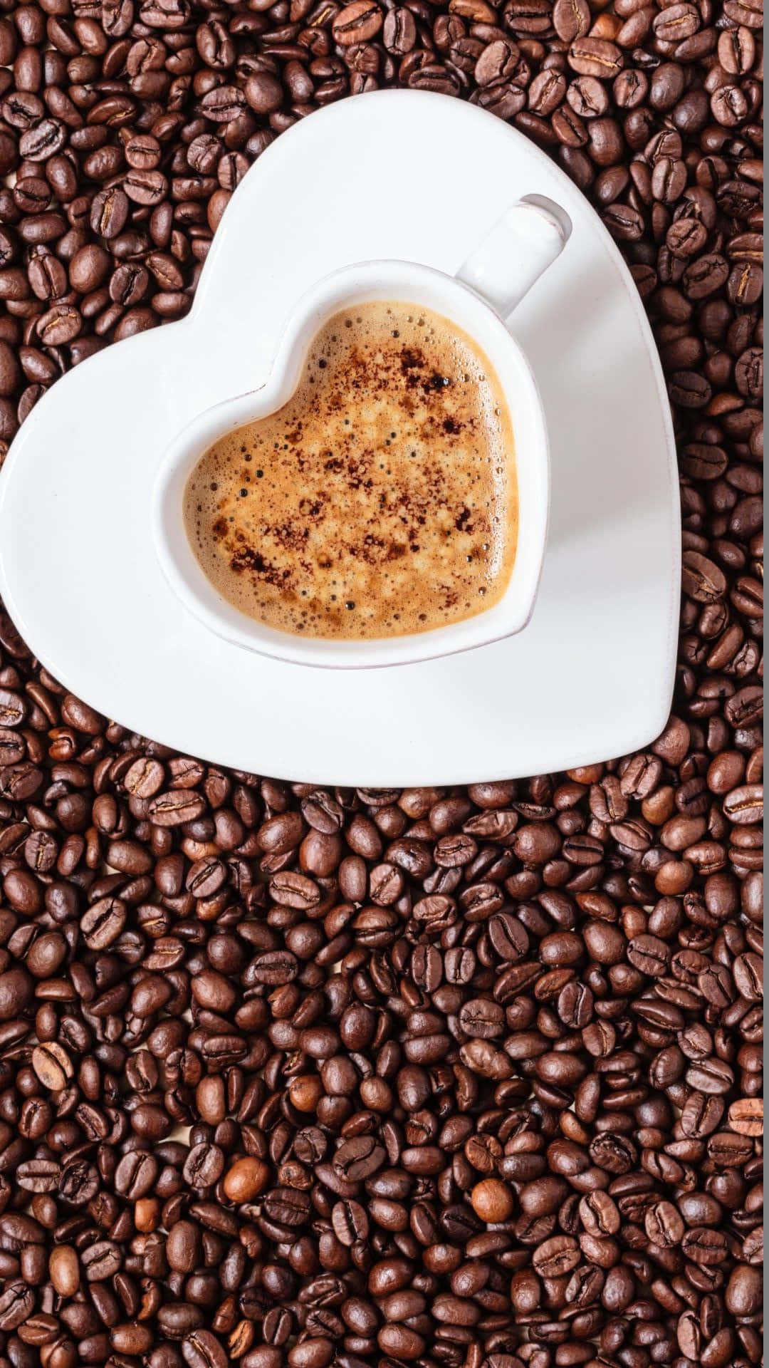 Coffee Aesthetic 桌布 壁紙 — 一個白色心形杯裝滿泡沫咖啡，放在深烘焙咖啡豆上。