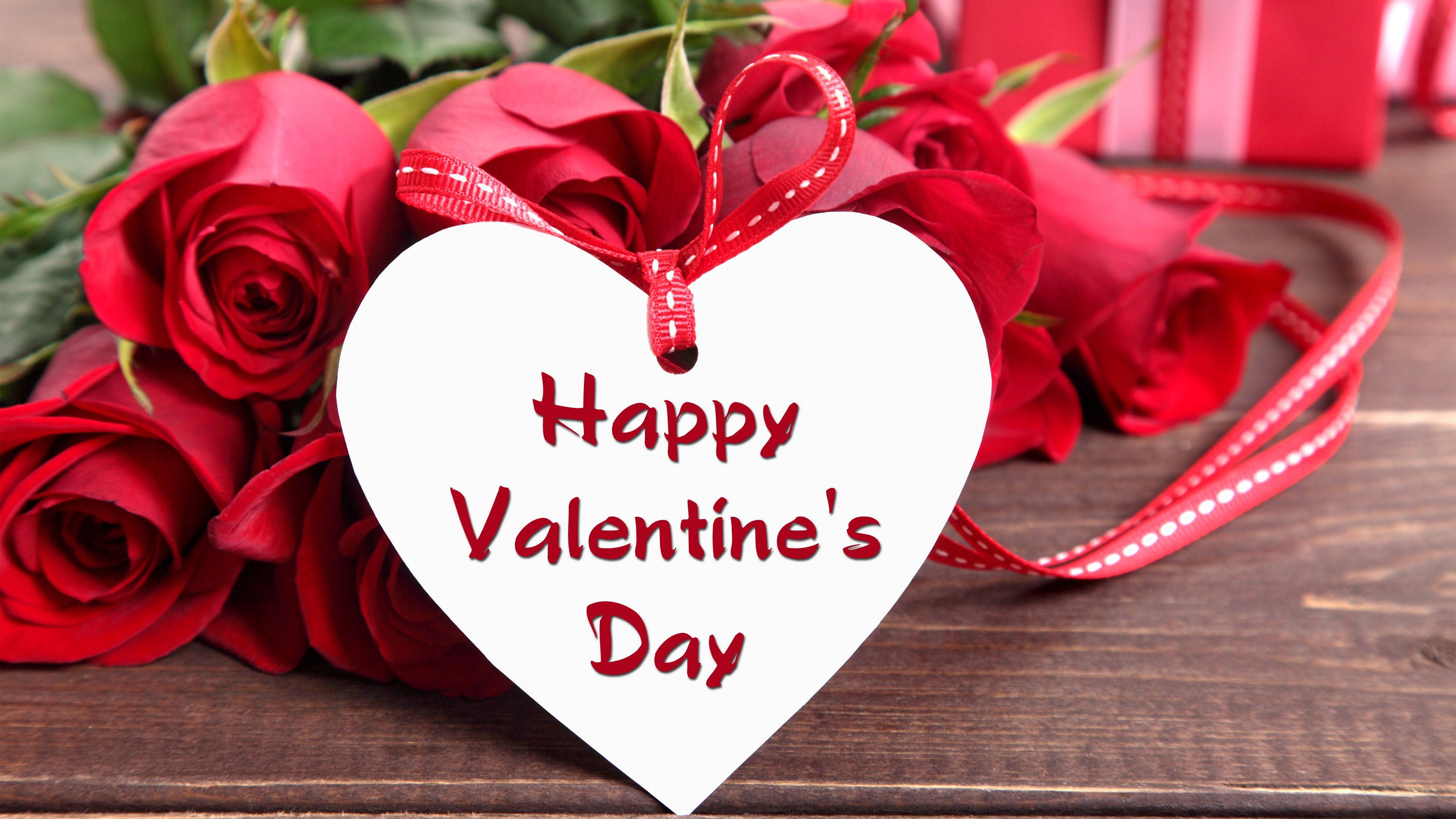 情人節 桌布 壁紙 — 一個白色心形標籤，上面有紅色草書文字寫著「Happy Valentine's Day」，用紅色絲帶綁著，在鮮豔的紅玫瑰中，置於鄉村風格的木質表面上。