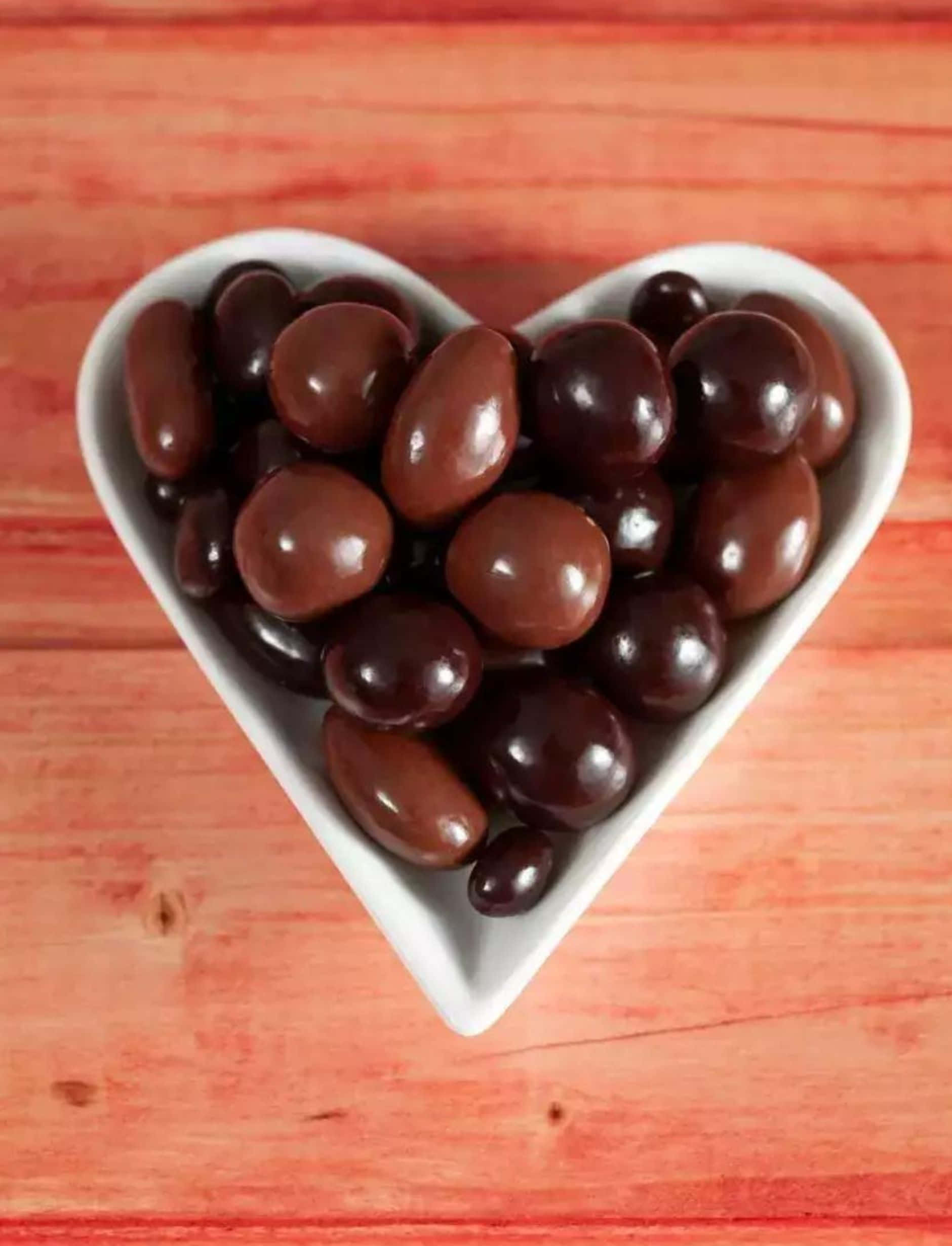 Dark Chocolate 桌布 壁紙 — 一個白色心形碗裝滿了光澤的深色巧克力糖果，放在暖色調的木質表面上。