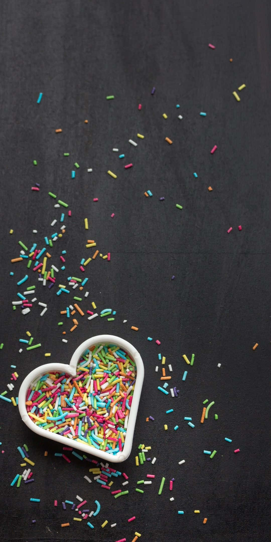 Sprinkles 桌布 壁紙 — 一個白色心形碗裝滿彩色糖粒，灑落在深色紋理表面上。