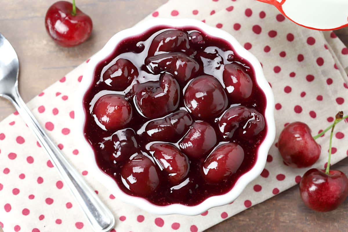 Red Cherries 桌布 壁紙 — 一個白色扇貝邊碗，盛滿了糖漿中閃亮的深紅色櫻桃，放在紅色圓點餐巾上，旁邊有新鮮櫻桃和湯匙。