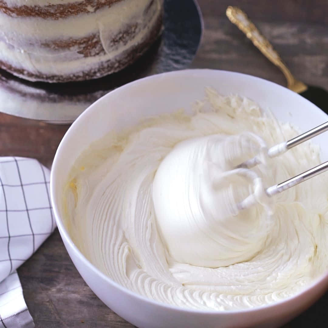 Buttercream 桌布 壁紙 — 一個白色攪拌碗裝滿蓬鬆的鮮奶油，正用電動攪拌器攪拌，背景有層層蛋糕。