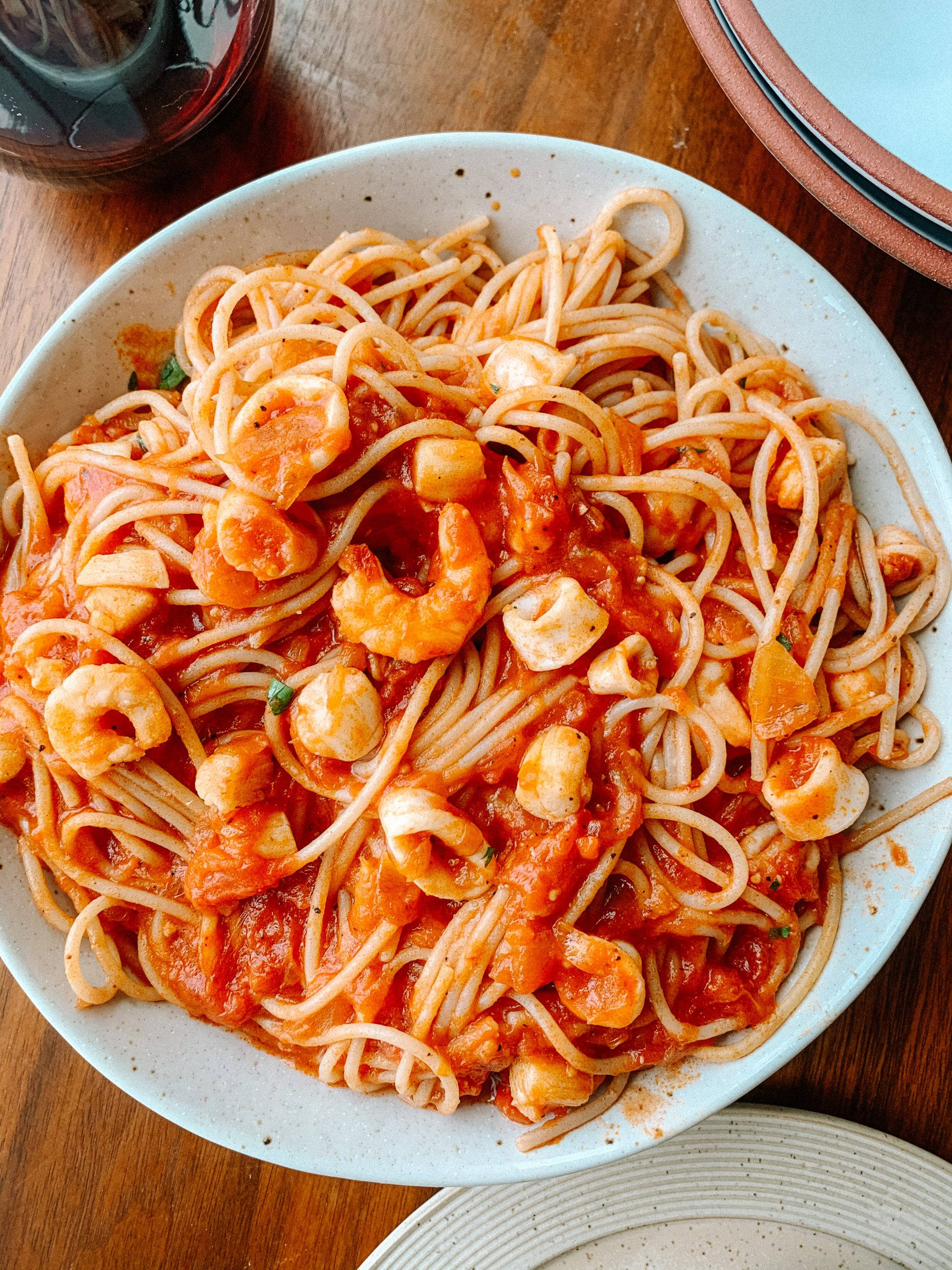 Seafood 桌布 壁紙 — 一個白色斑點碗的特寫，裡面裝滿了義大利麵，配上濃郁的番茄醬，上面放著蝦和蛤蜊，放在木桌上。
