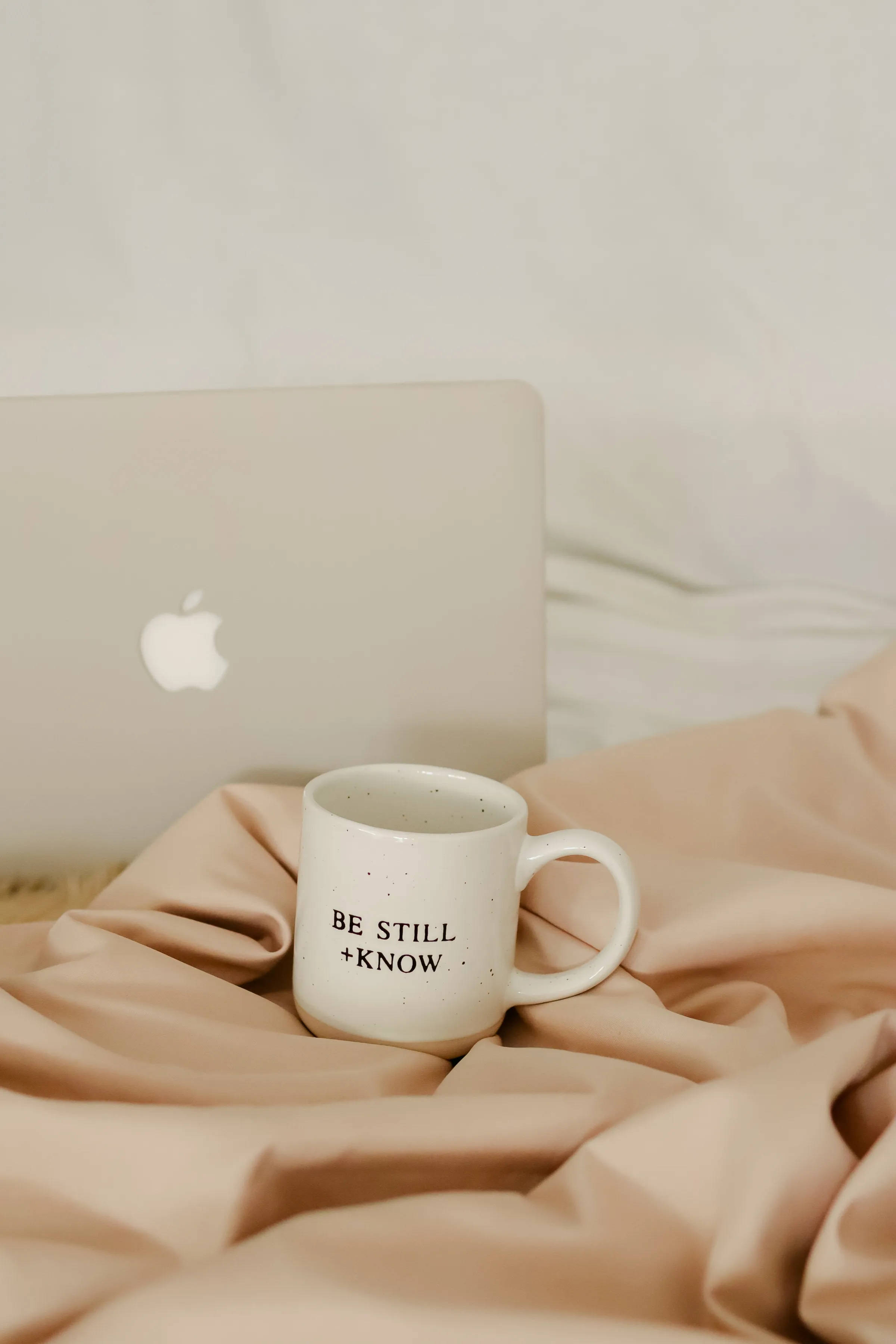 Coffee Aesthetic 桌布 壁紙 — 一個白色斑點馬克杯，上面寫著「BE STILL + KNOW」，放在床上柔軟的米色布料上，旁邊有一台銀色筆記型電腦。