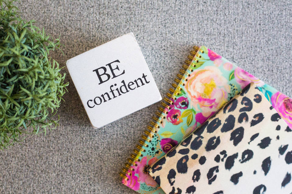 勵志桌面 桌布 壁紙 — 一個白色方塊，上面有粗體黑色文字寫著「BE confident」，旁邊放著一本花卉圖案筆記本和一本豹紋圖案筆記本，放在有紋理的灰色表面上。