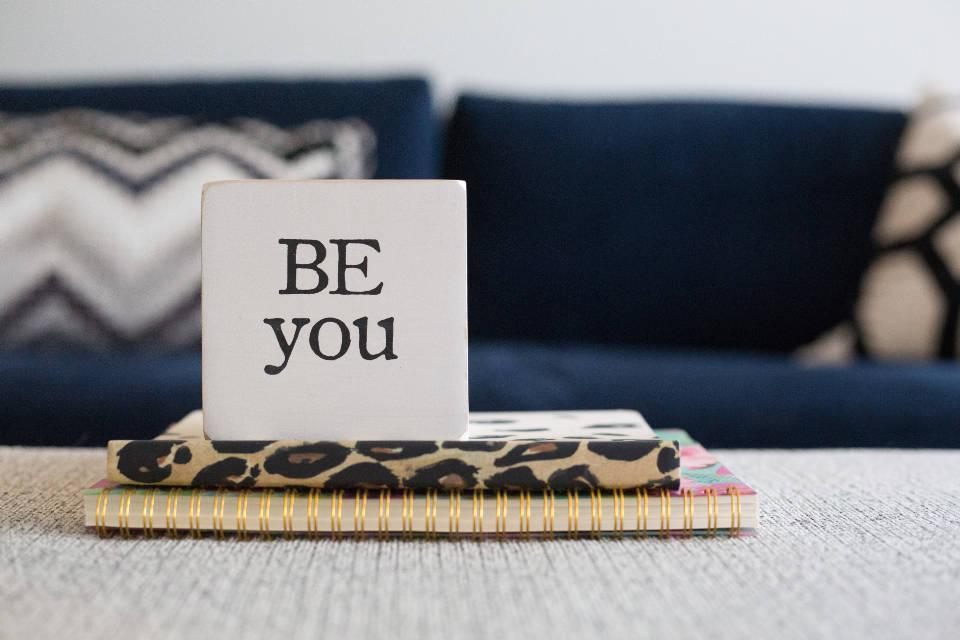 勵志桌面 桌布 壁紙 — 一個白色方塊上寫著「BE you」，放在有圖案的筆記本上，置於有紋理的桌子上，前面是海軍藍沙發。