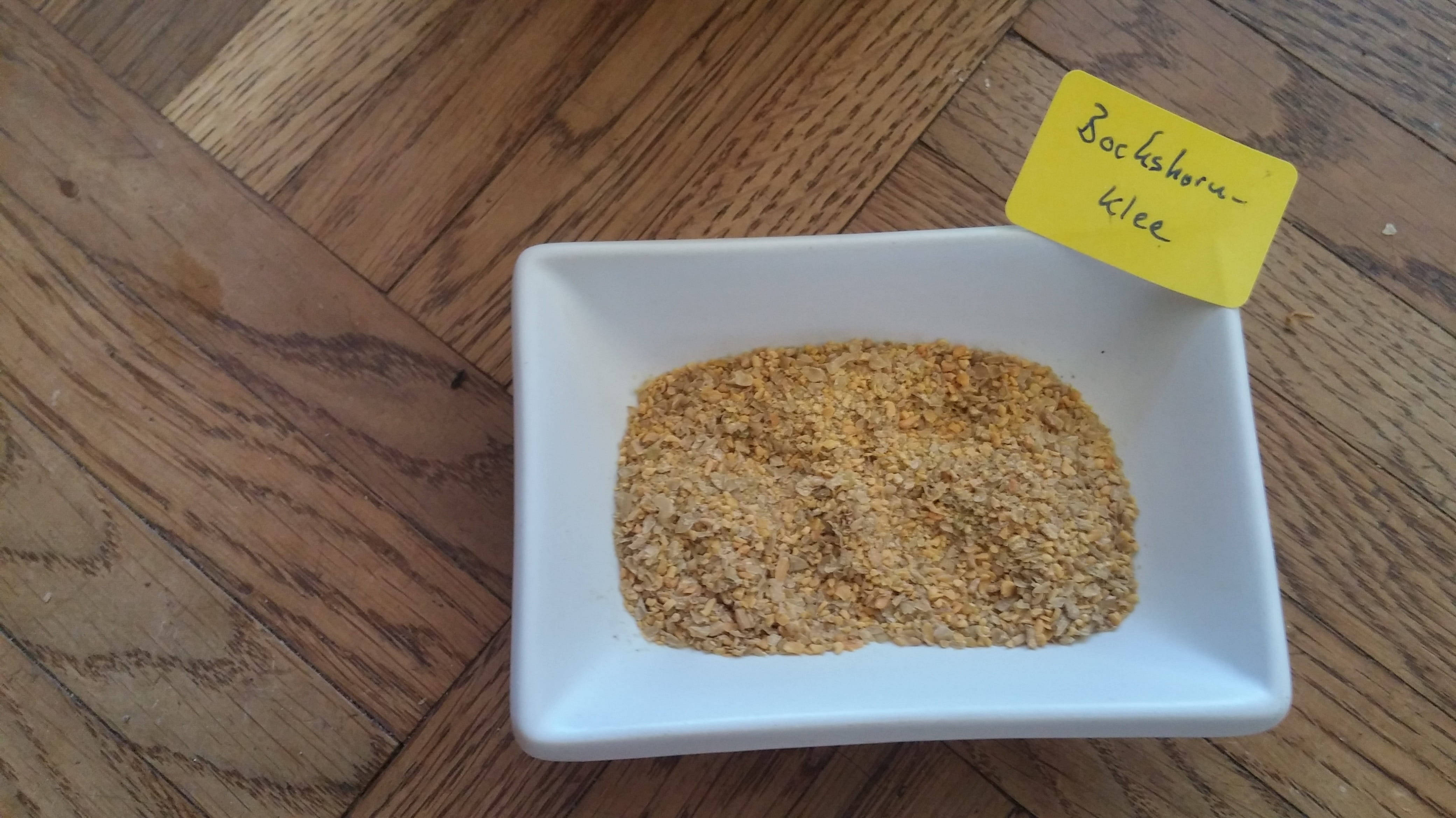 烹飪 桌布 壁紙 — 一個白色方形盤子盛裝著金黃色的碎Bockshorn-Klee種子，放在溫暖的木地板上，附有黃色標籤。