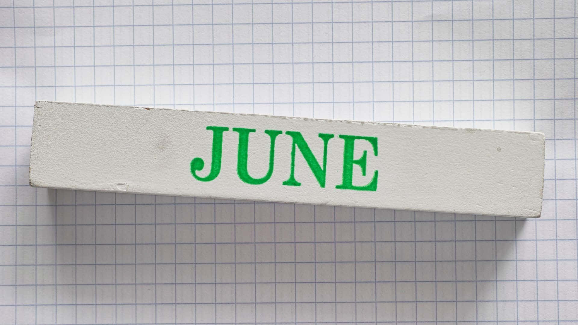 六月 桌布 壁紙 — 一個白色木塊上帶有綠色 'JUNE' 字樣，放在藍色方格紙上，光線明亮。