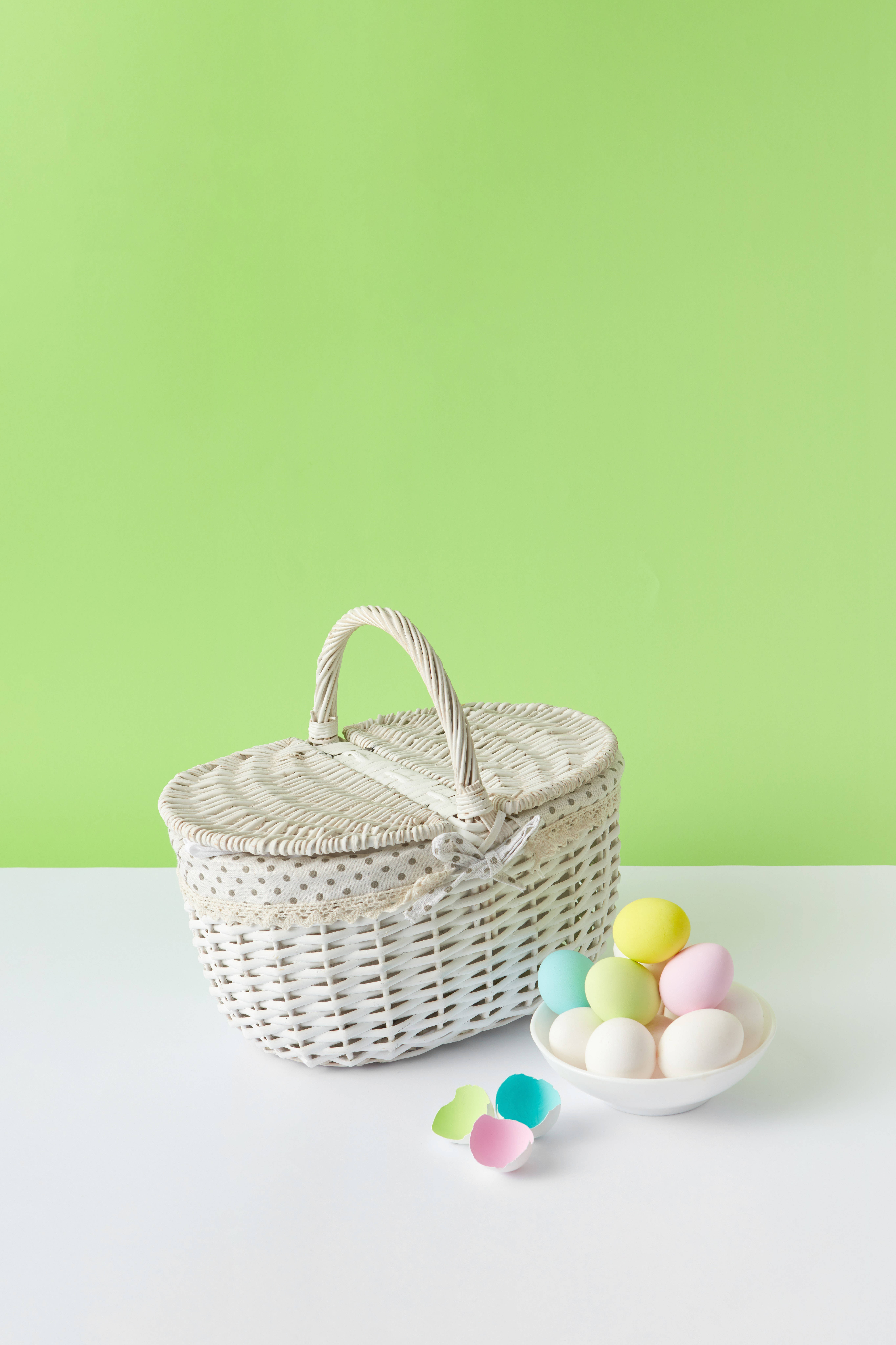 Cute Pastel Aesthetic 桌布 壁紙 — 一個白色柳條復活節籃子，帶有圓點飾邊，旁邊放著一碗粉彩雞蛋和散落的蛋殼半邊，背景是明亮的綠色牆壁。