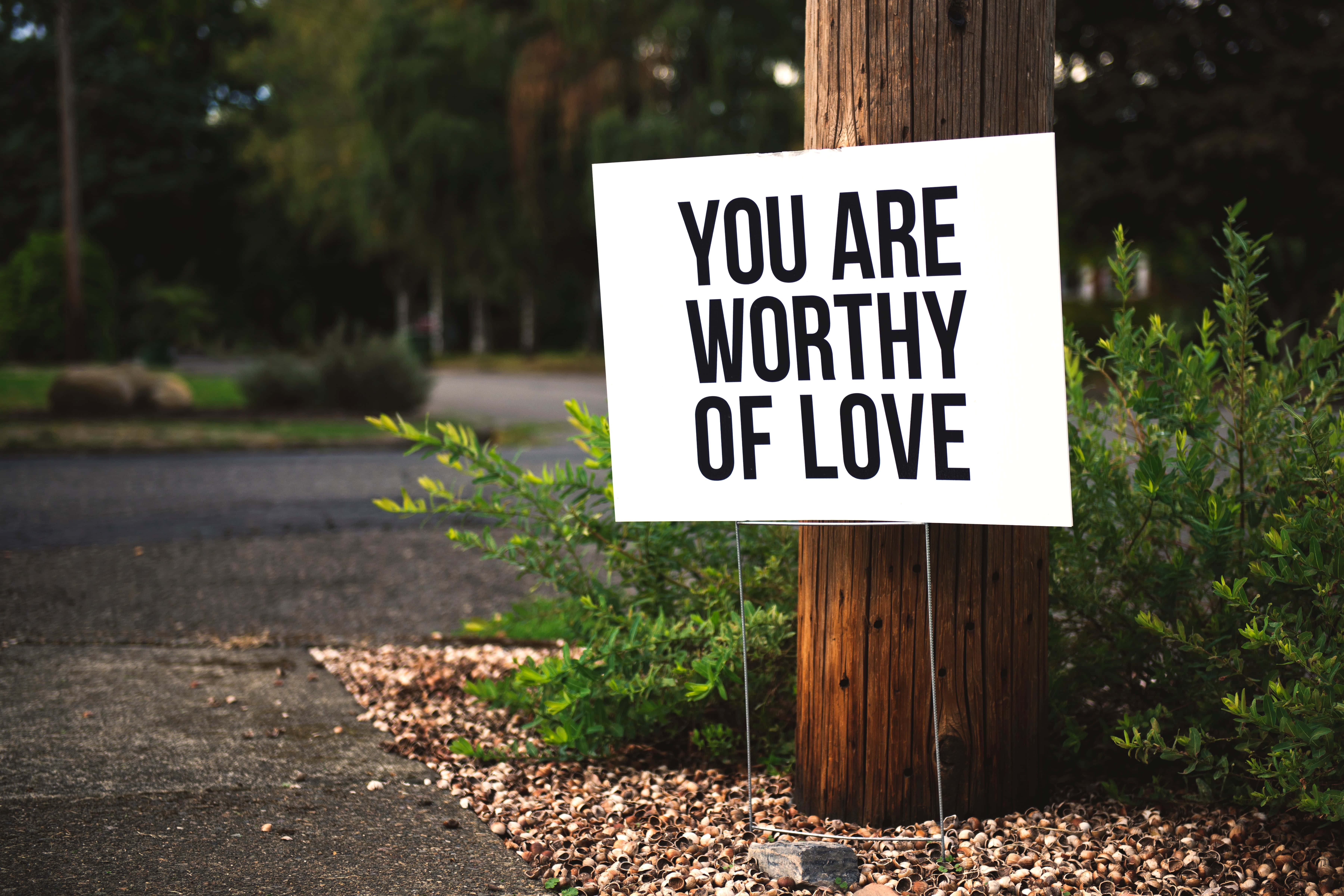 桌布 壁紙 — 一個白色標牌，上面寫著「YOU ARE WORTHY OF LOVE」，豎立在碎石路邊花園的木柱旁。