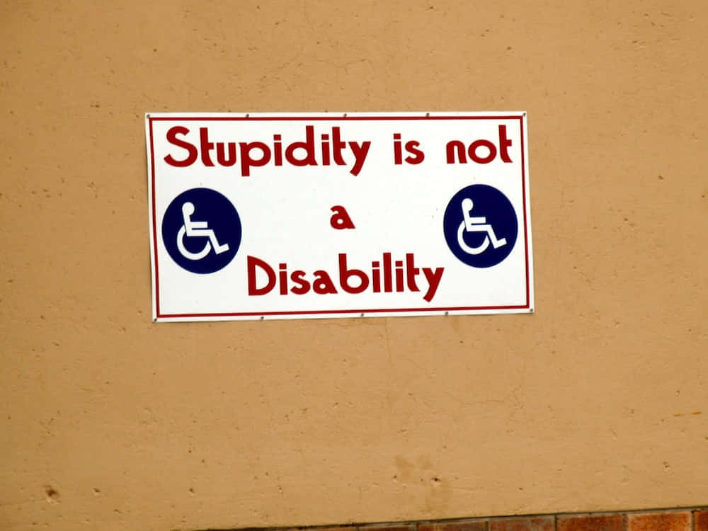 Stupid 桌布 壁紙 — 一個白色標牌，上面有紅色文字寫著「Stupidity is not a Disability」，兩側有兩個藍色輪椅符號，安裝在米色牆上。