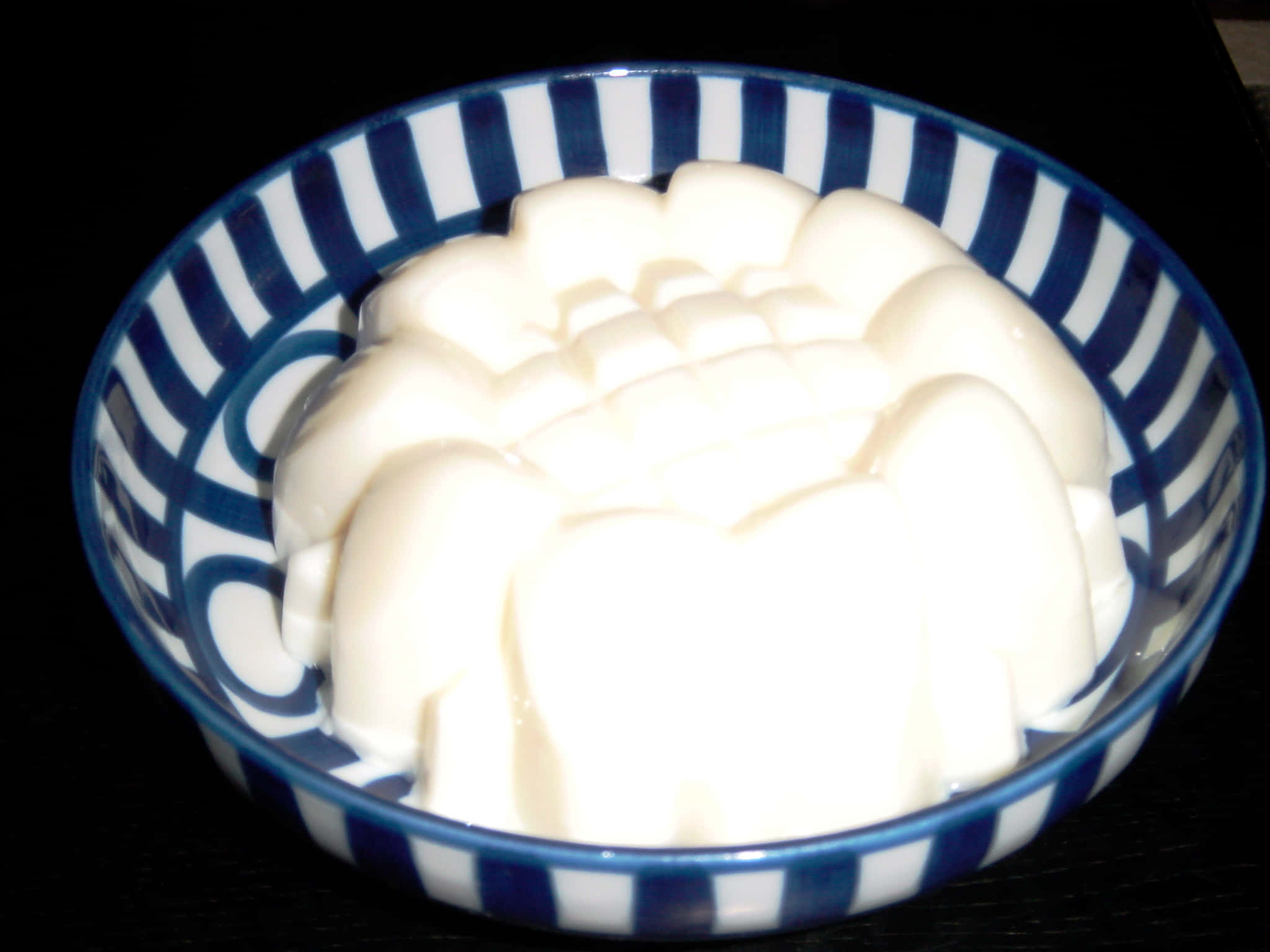 Blancmange 桌布 壁紙 — 一個白色模製果凍甜點，邊緣呈扇貝形，放在一個藍白條紋陶瓷碗中，背景為深色。