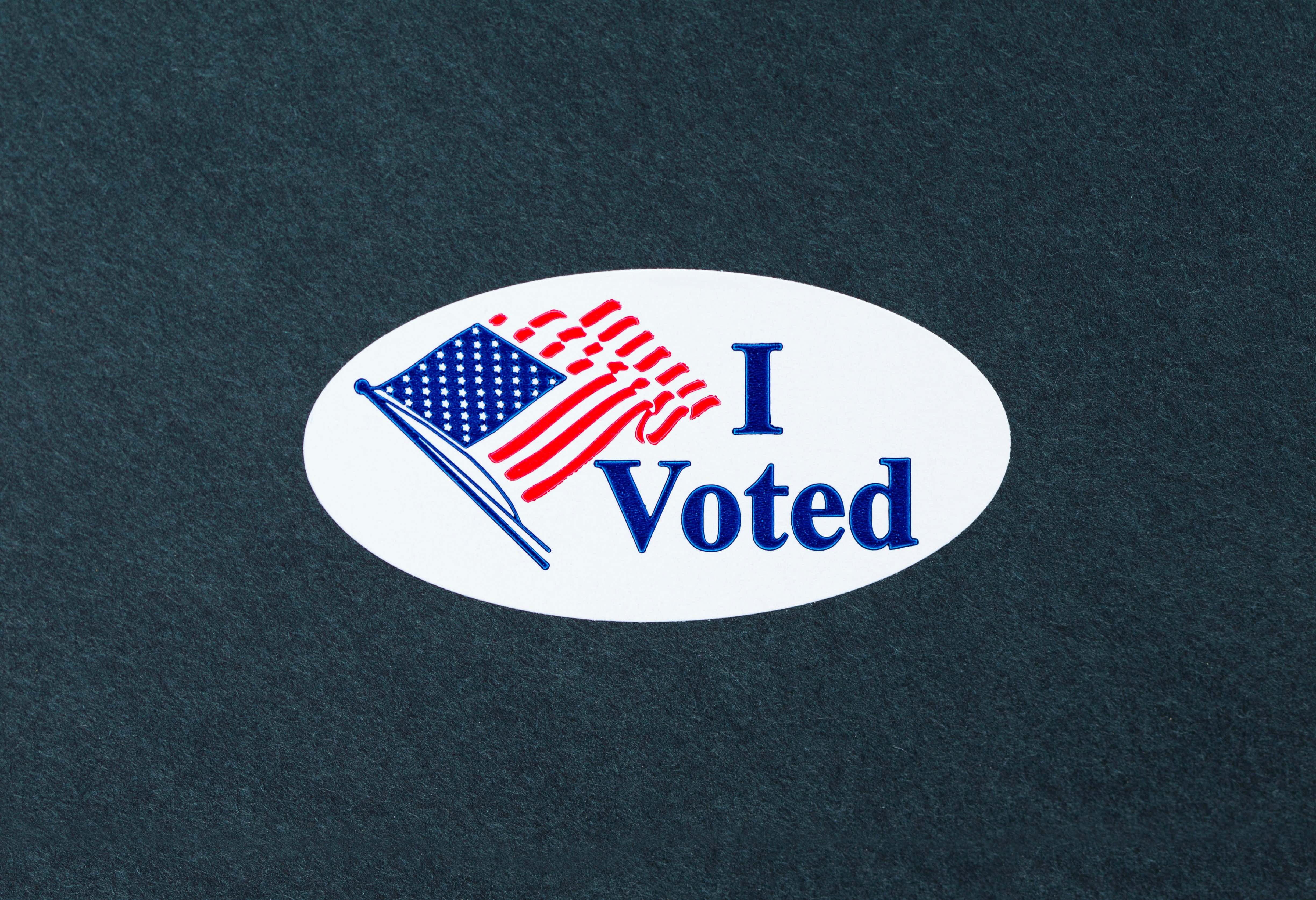 選舉 桌布 壁紙 — 一個白色橢圓形貼紙，上面有藍色文字寫著「I Voted」，以及一個風格化的美國國旗圖案，位於有紋理的深藍色布料上。