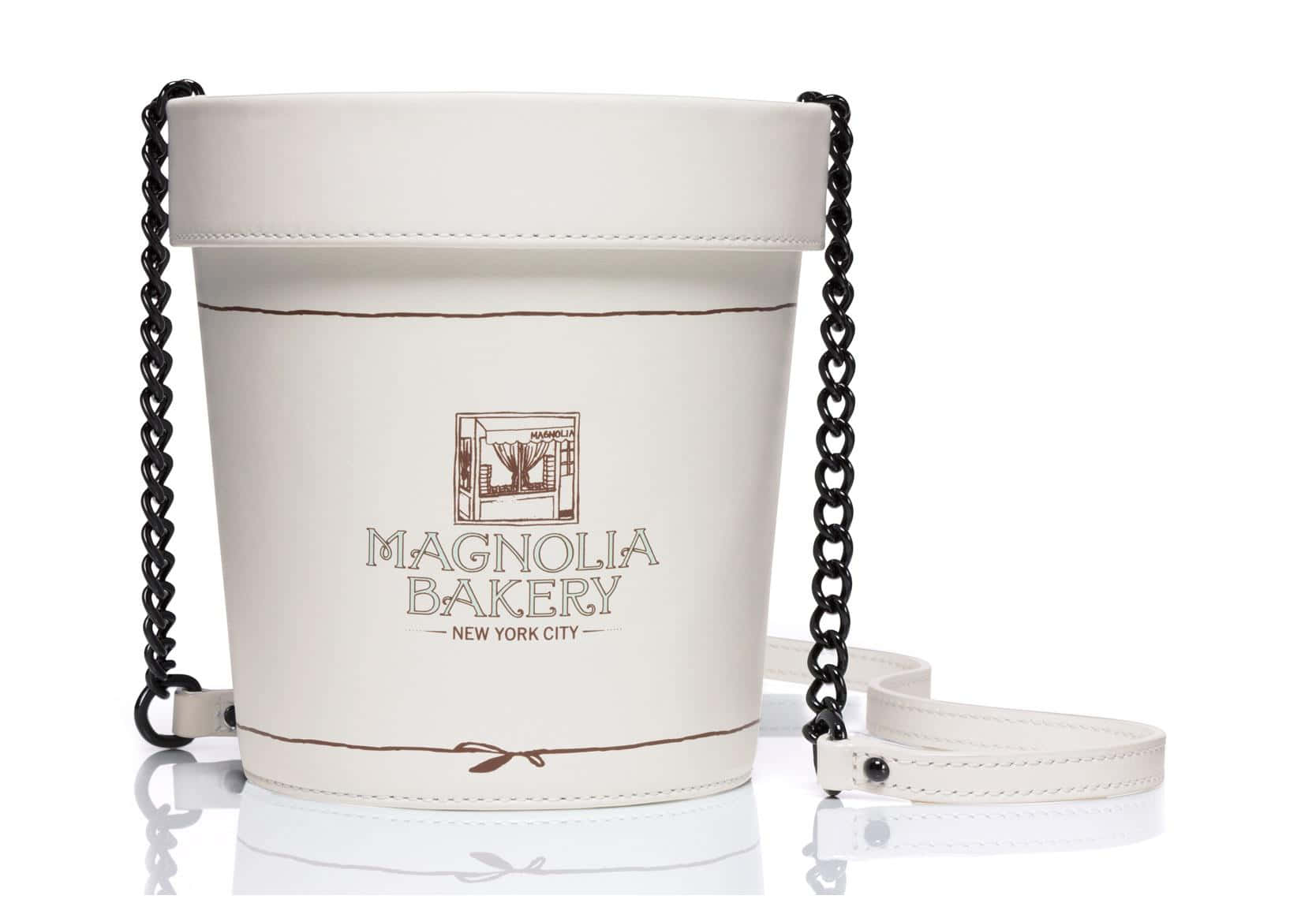 Kate Spade 桌布 壁紙 — 一個白色水桶形手提包，配有黑色鏈條肩帶和 Magnolia Bakery 品牌標誌，放置在反光表面上。