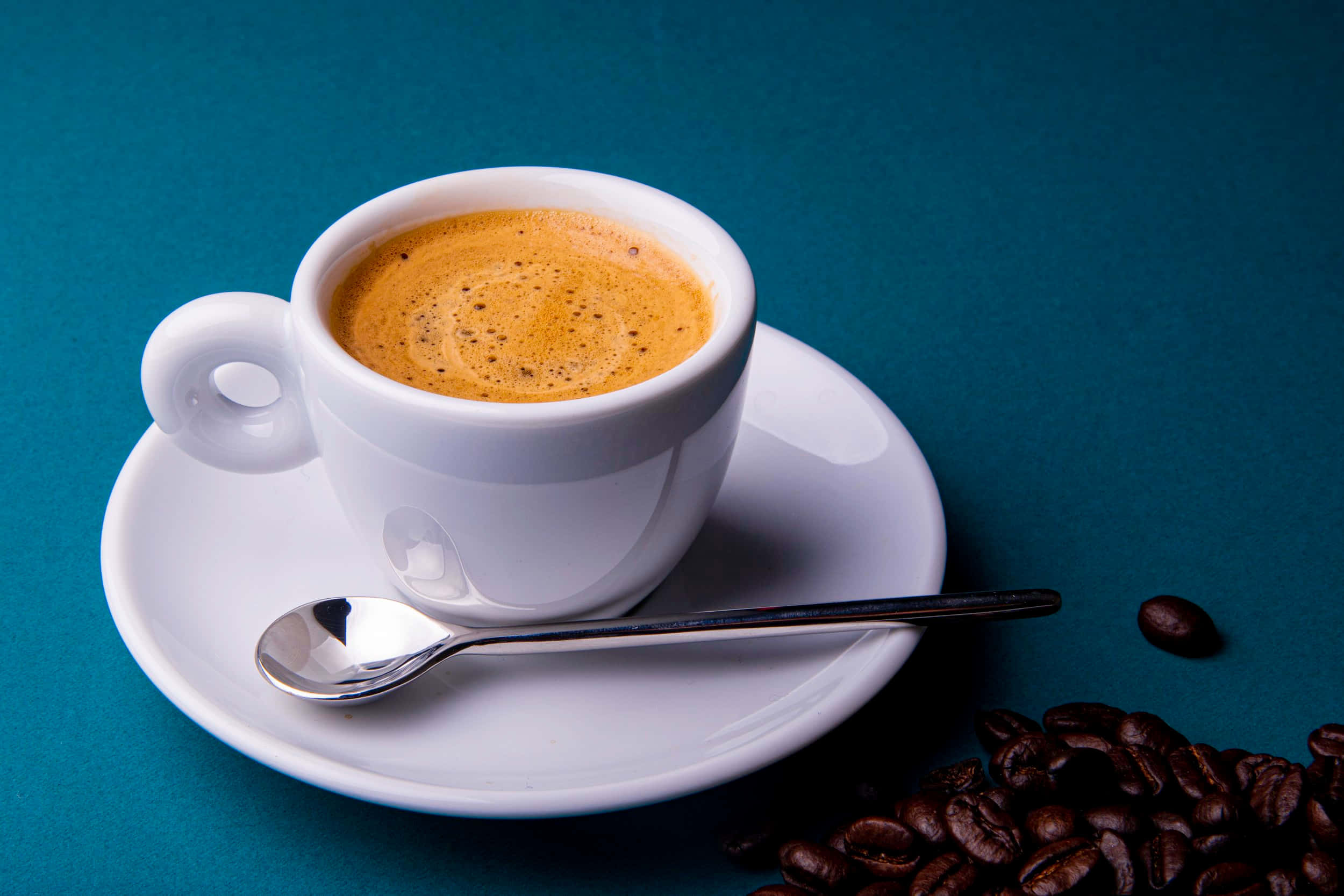 Milk And Coffee 桌布 壁紙 — 一個白色濃縮咖啡杯盛載著泡沫咖啡，放在碟子上，附有湯匙和散落的烘焙咖啡豆，背景為青綠色。