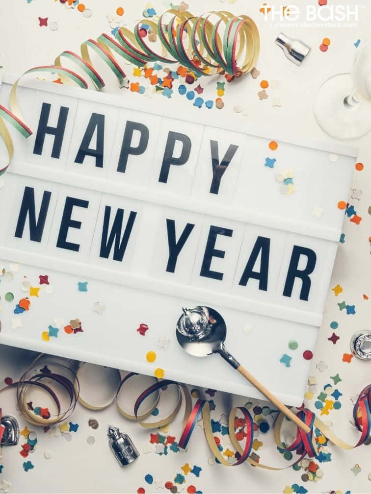 節日 桌布 壁紙 — 一個白色燈箱顯示「HAPPY NEW YEAR」，周圍環繞著彩色紙屑絲帶和香檳瓶，放在明亮的表面上。