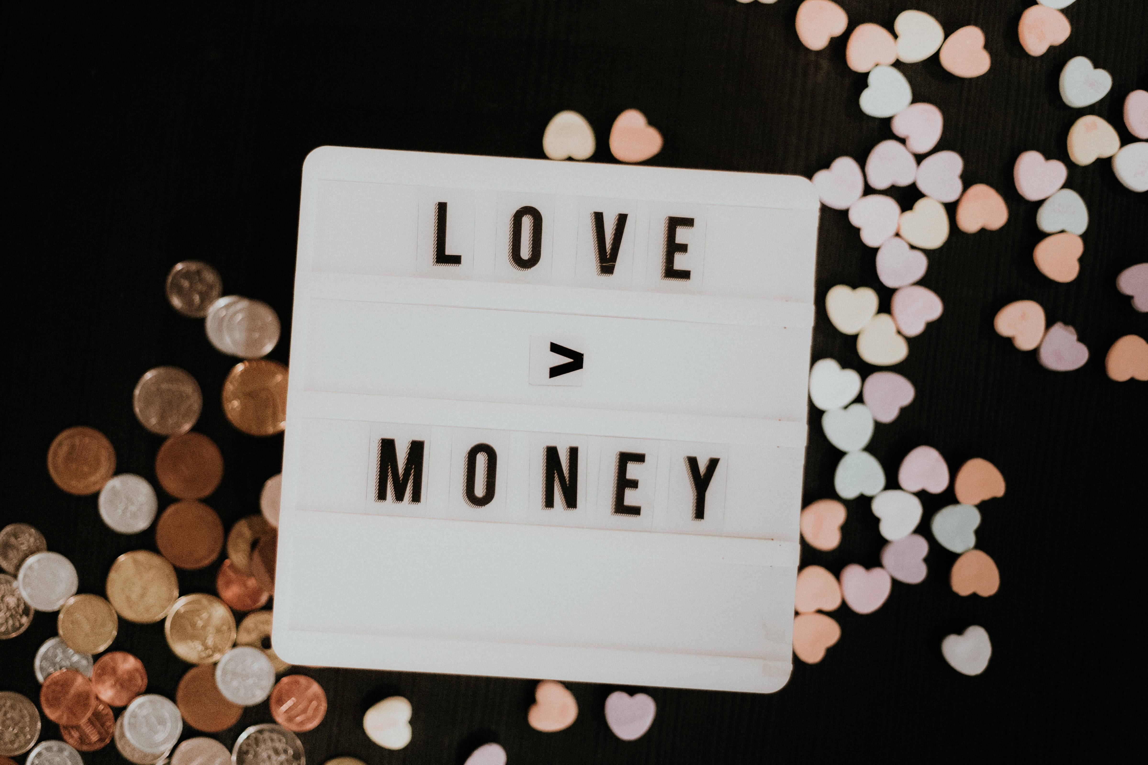 我愛你 桌布 壁紙 — 一個白色燈箱顯示「LOVE > MONEY」，周圍有硬幣和粉彩心形紙屑在黑色表面上。