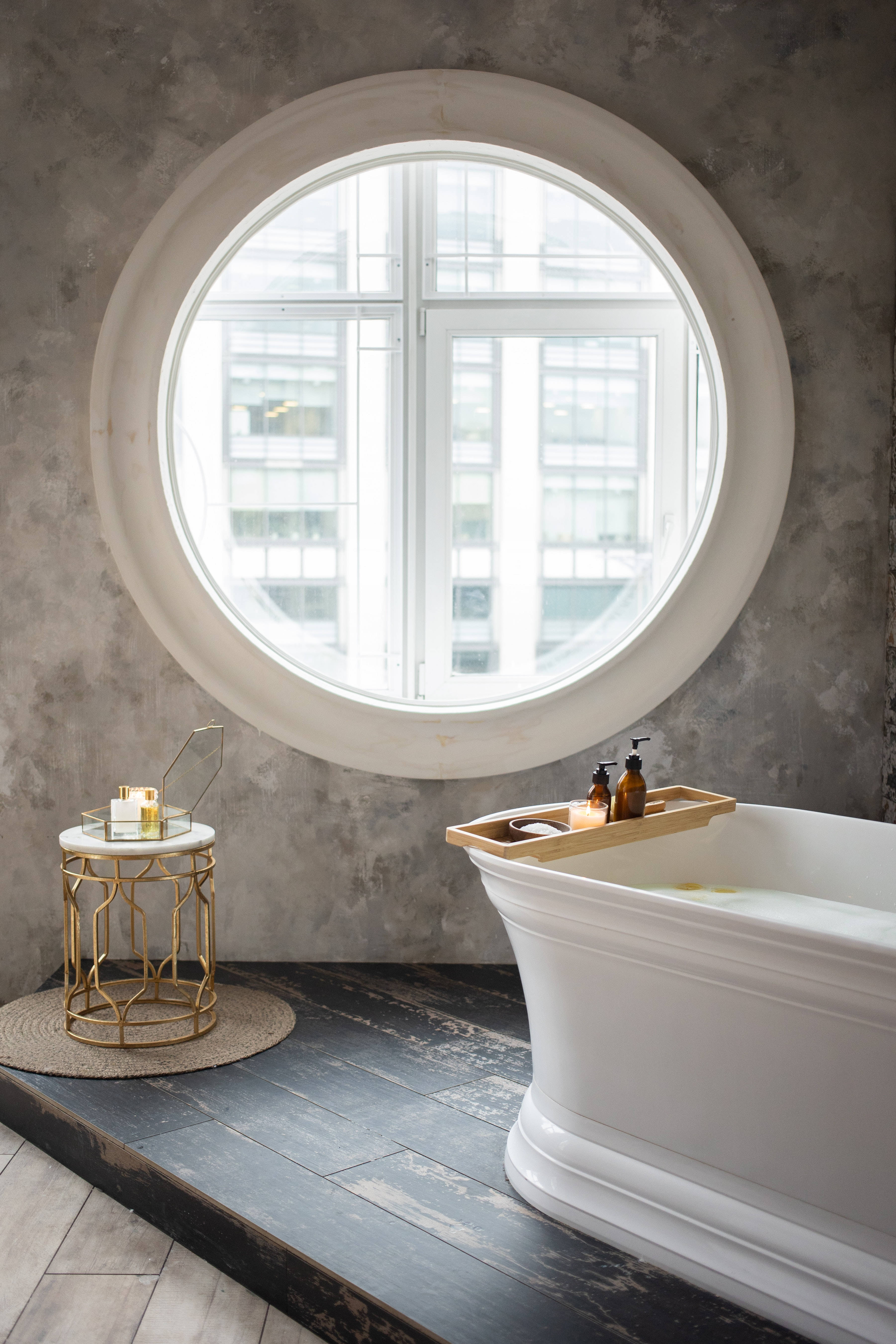 Bathtub 桌布 壁紙 — 一個白色獨立式浴缸位於一個大型圓窗下方，窗外可俯瞰城市建築，浴室採用現代風格，配有紋理灰色牆壁和深色木地板。